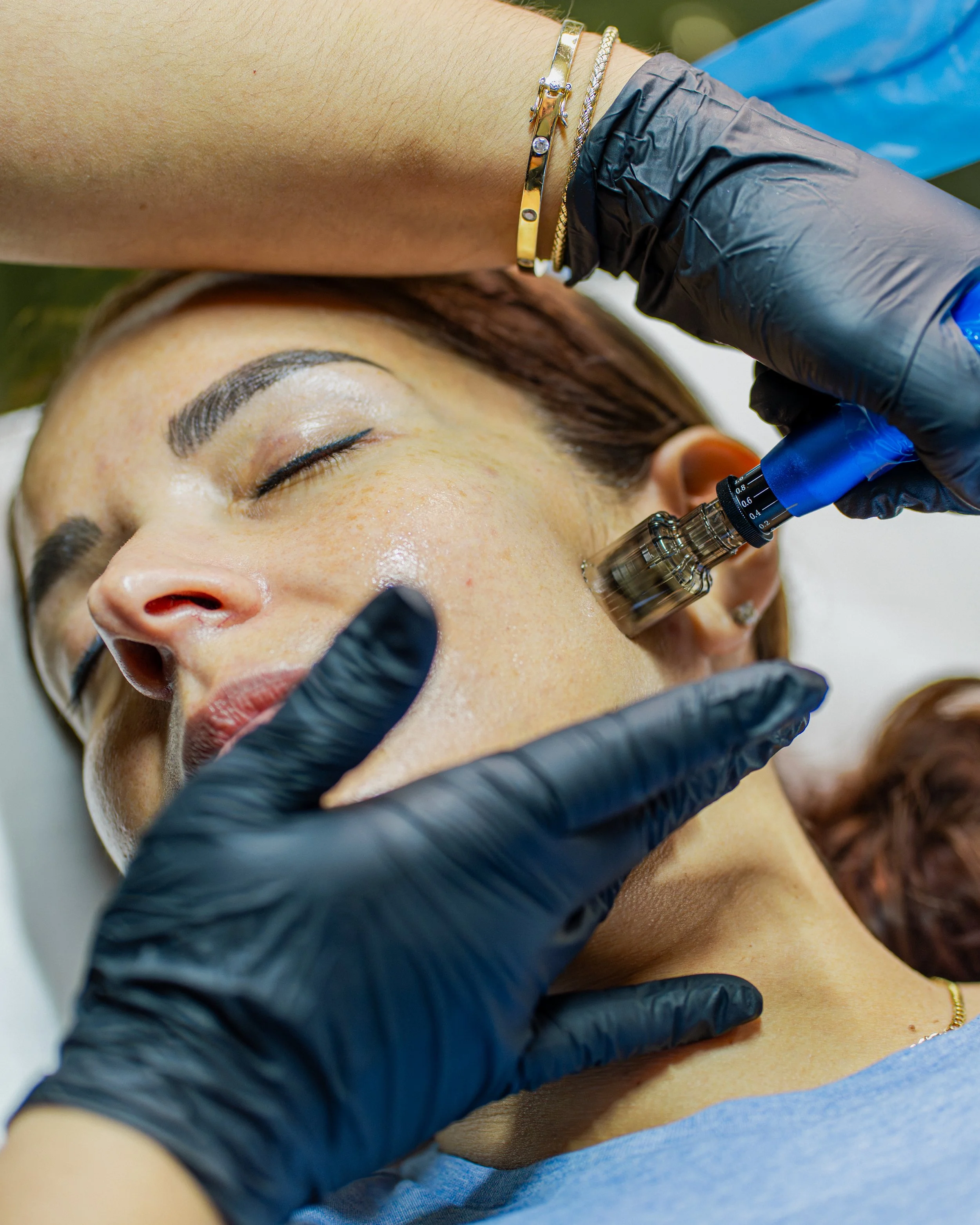 A woman receives a facial treatment with a jade roller, lying on a treatment bed covered with a white towel, her eyes closed, wearing a headband, with small leaves on her face.