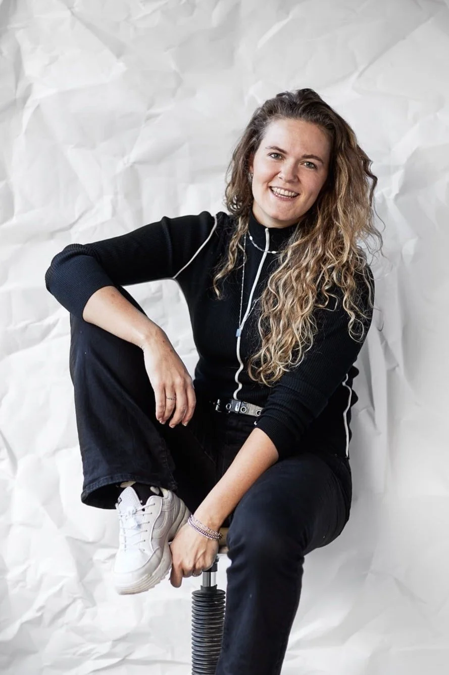 Vrouw met lang, krullend haar die glimlachend op een stoel zit, tegen een witte, gekreukte achtergrond.