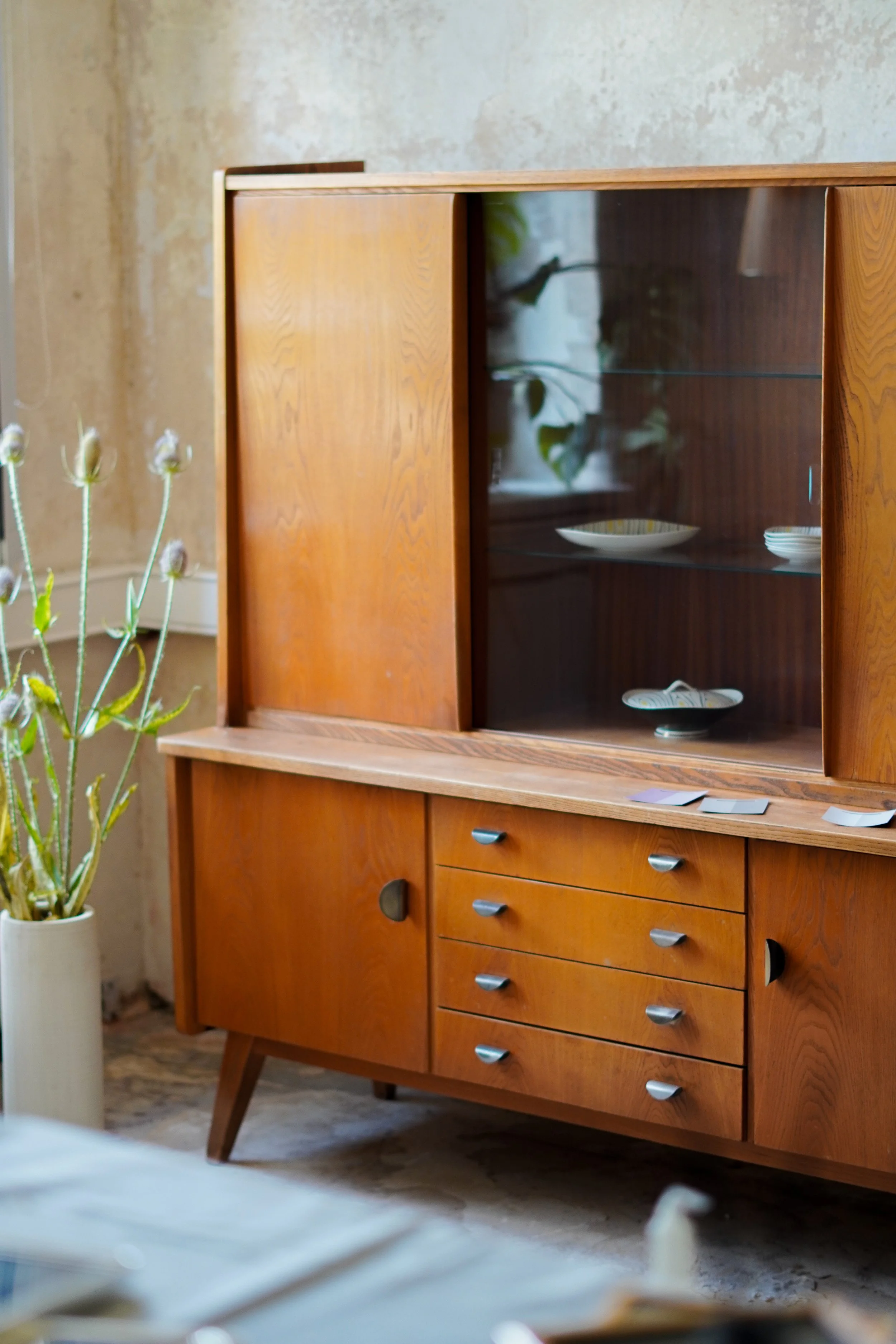 Ein alter Holzschrank mit Glastüren, darin Geschirr, neben einem weißen Pflanzgefäß mit grünen und weißen Pflanzen.