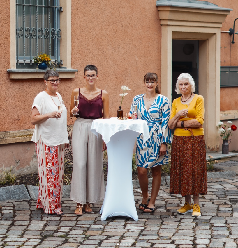 Vier Frauen stehen im Hintergrund einer Hauswand auf einem gepflasterten Hof, die eine Cocktailparty oder Feier veranstalten. Sie halten Gläser mit Getränken in der Hand. Es gibt eine hohe Stehtafel mit einer weißen Tischdecke, auf der eine Flasche und eine Vase mit weißen Blumen stehen. Die Frauen sind in sommerlicher Kleidung und erscheinen fröhlich.