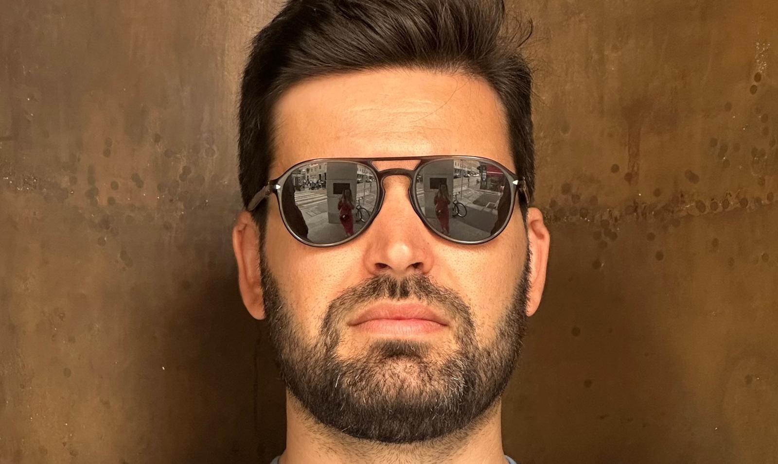 A man wearing dark sunglasses with a reflection of an outdoor city scene, including a person taking the photo, in the lenses, and a textured brown wall in the background.