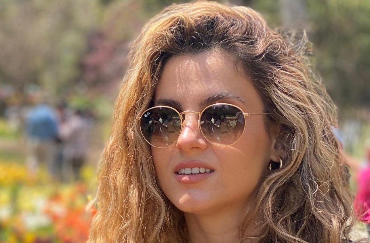 A woman with curly hair wearing round sunglasses and earrings outdoors, with trees and people in the background.