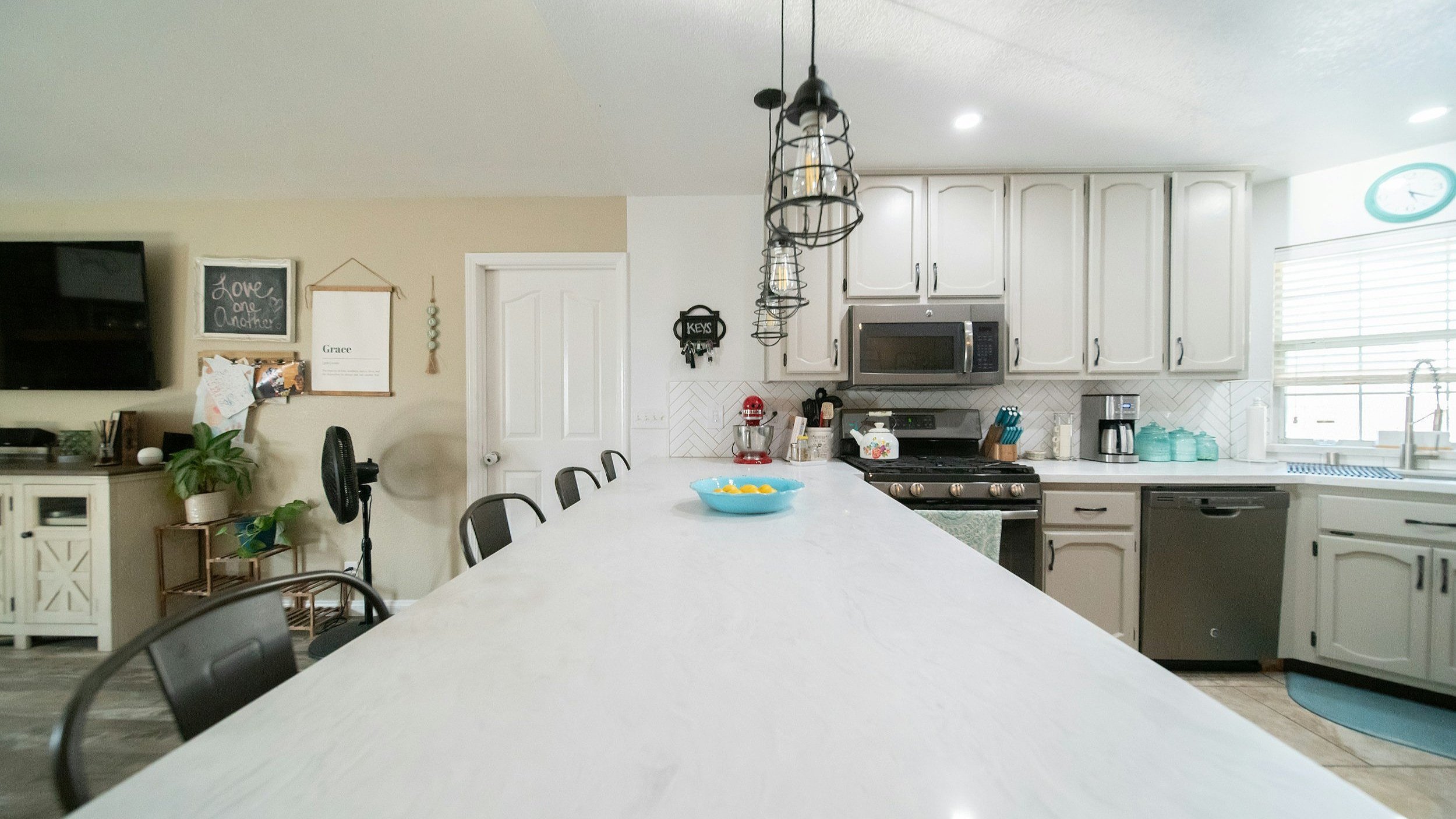 Modern kitchen with white cabinets, black appliances, and a large white island. Pendant lights hang above the island. There is a microwave, coffee maker, and utensils on the counters. A window with blinds lets in natural light. A wall clock and decor