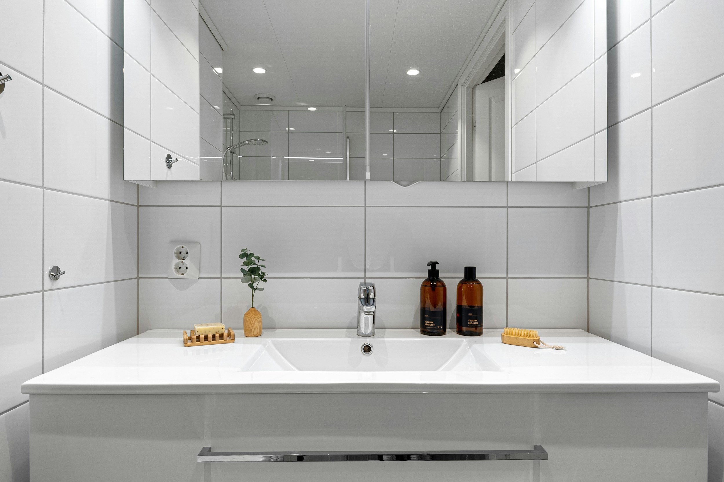 Bathroom sink with soap, plant, and amber bottles, mirror reflecting shower area.