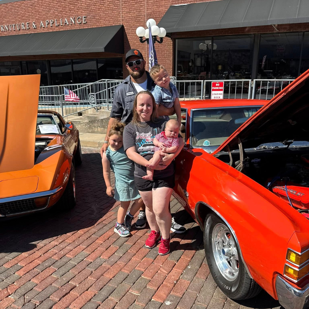 A family standing next to a classic orange car with its hood open at a car show, with another vintage car partly visible on the left. The family consists of a man, woman, and three children, smiling for the photo, outside a brick building with a sign that reads 'Furniture & Appliance'.