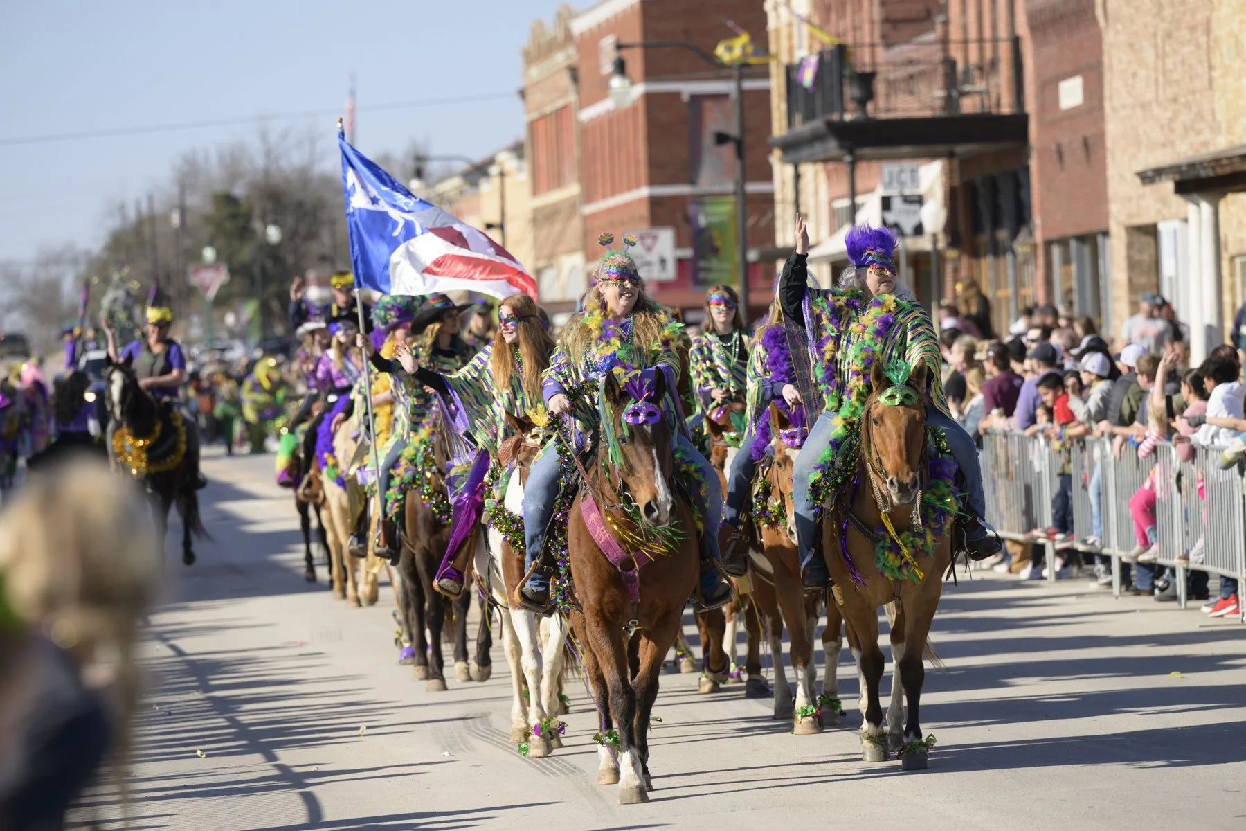 People participating in a parade, dressed in colorful costumes, riding horses decorated with Mardi Gras beads and masks, with spectators watching on both sides of the street.