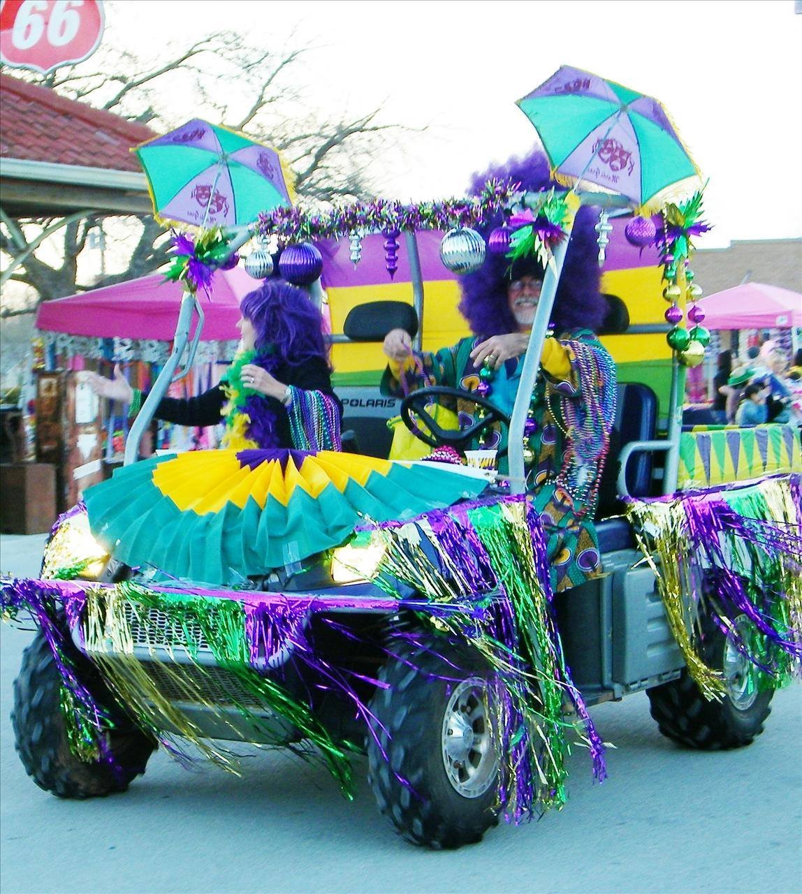 A decorated parade vehicle with two people dressed in colorful costumes. They are wearing purple wigs, sunglasses, and beads, and are surrounded by Mardi Gras-themed decorations with purple, green, and gold colors, including umbrellas, ornaments, and streamers.