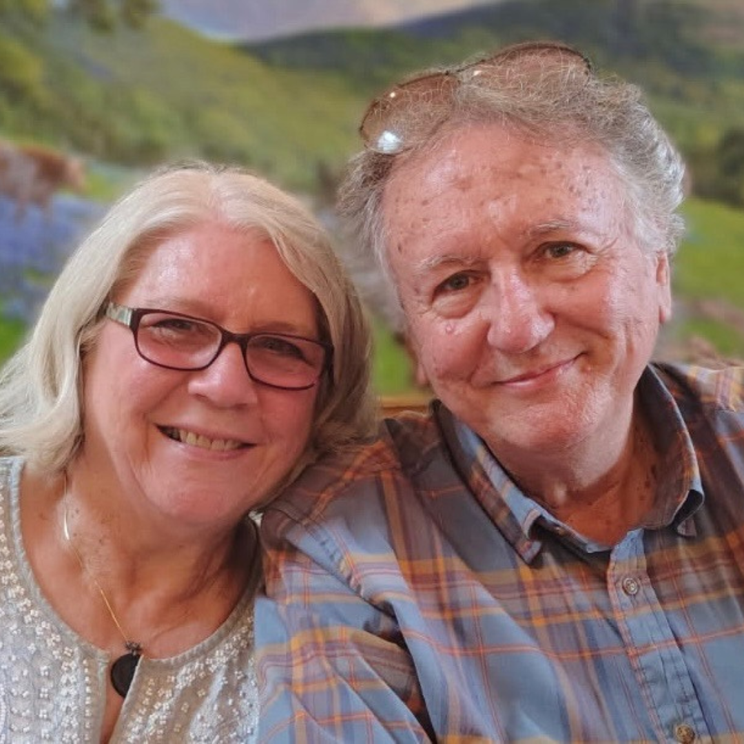 A smiling elderly couple taking a selfie outdoors with a scenic landscape background.