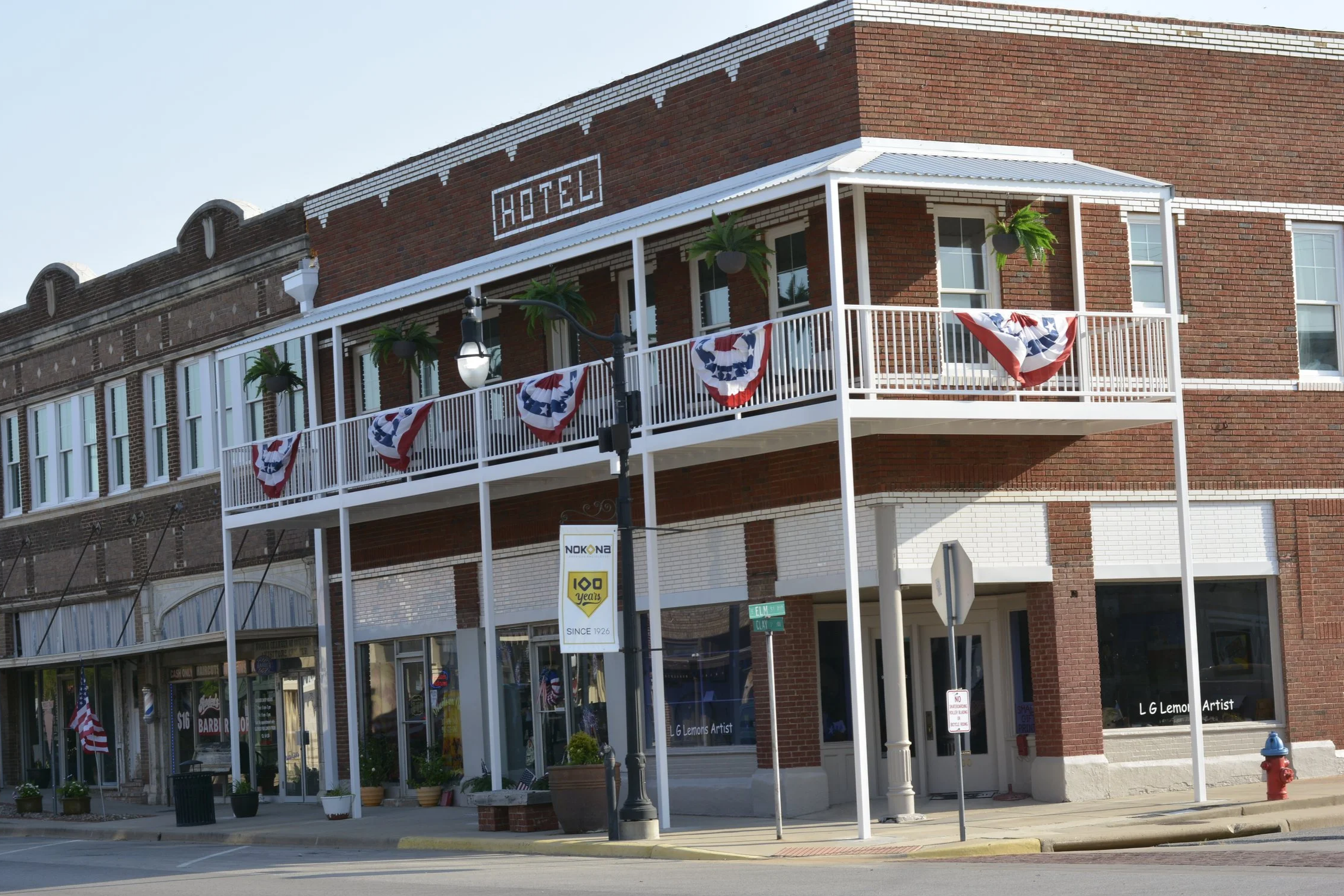 A brick building with a white balcony decorated with red, white, and blue bunting. There are potted plants hanging from the balcony and a streetlamp in front. The building has a sign that reads 'HOTEL'. Nocona  Texas Historical Hotel.