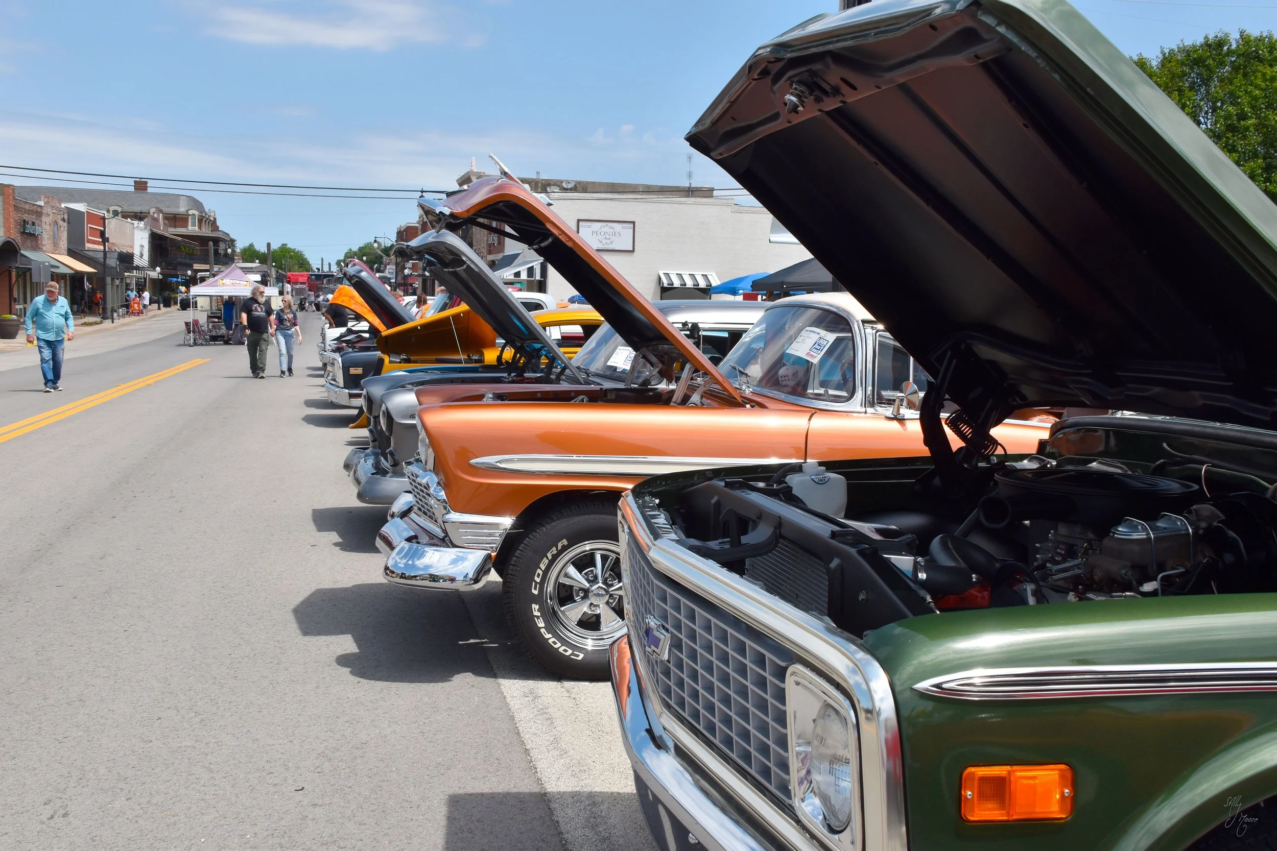 Line of vintage cars on display with hoods open at a street car show in a small town under a sunny sky.