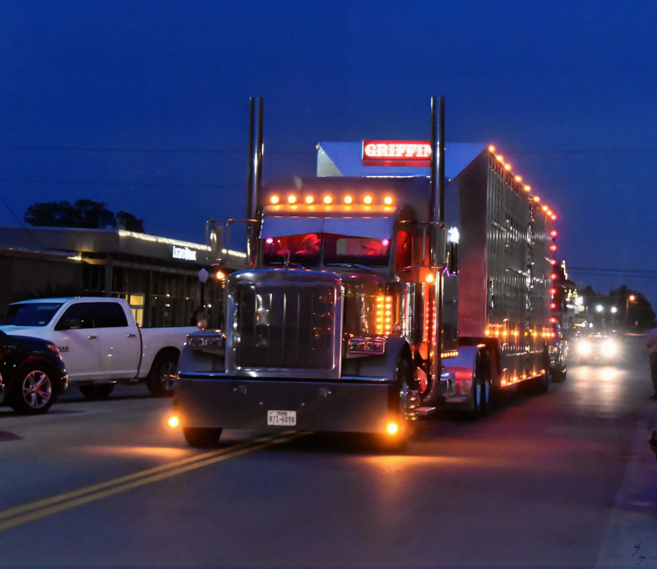 A large, shiny semi-truck with bright orange and yellow lights driving on a city street at night. There are other vehicles and a building with a sign in the background.