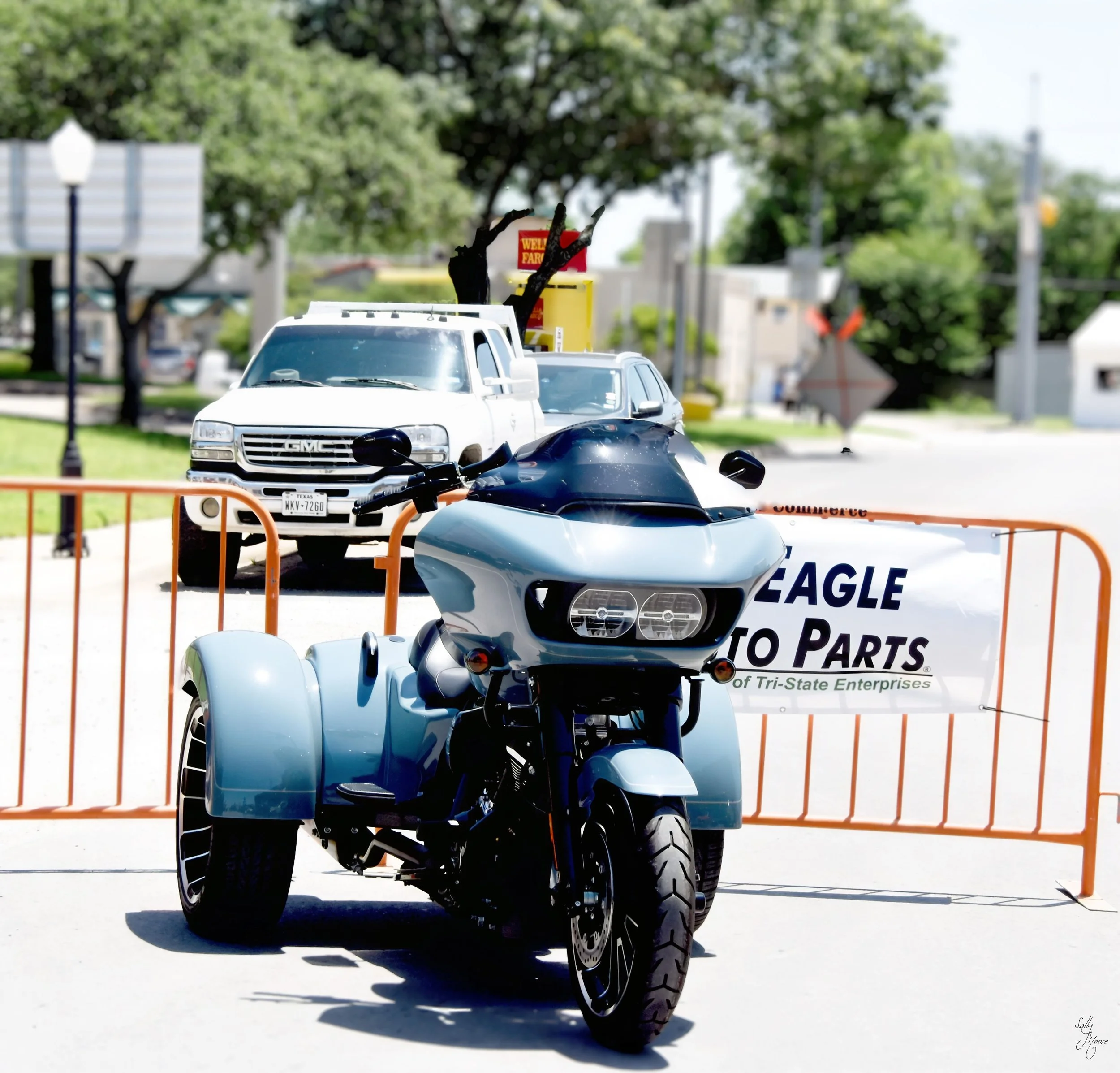 A white motorcycle with sidecar parked in front of an orange barricade with a sign that reads 'Eagle Auto Parts.' Behind the barricade, a white truck and a black car are visible. The background features trees, street signs, and a sunny outdoor setting.