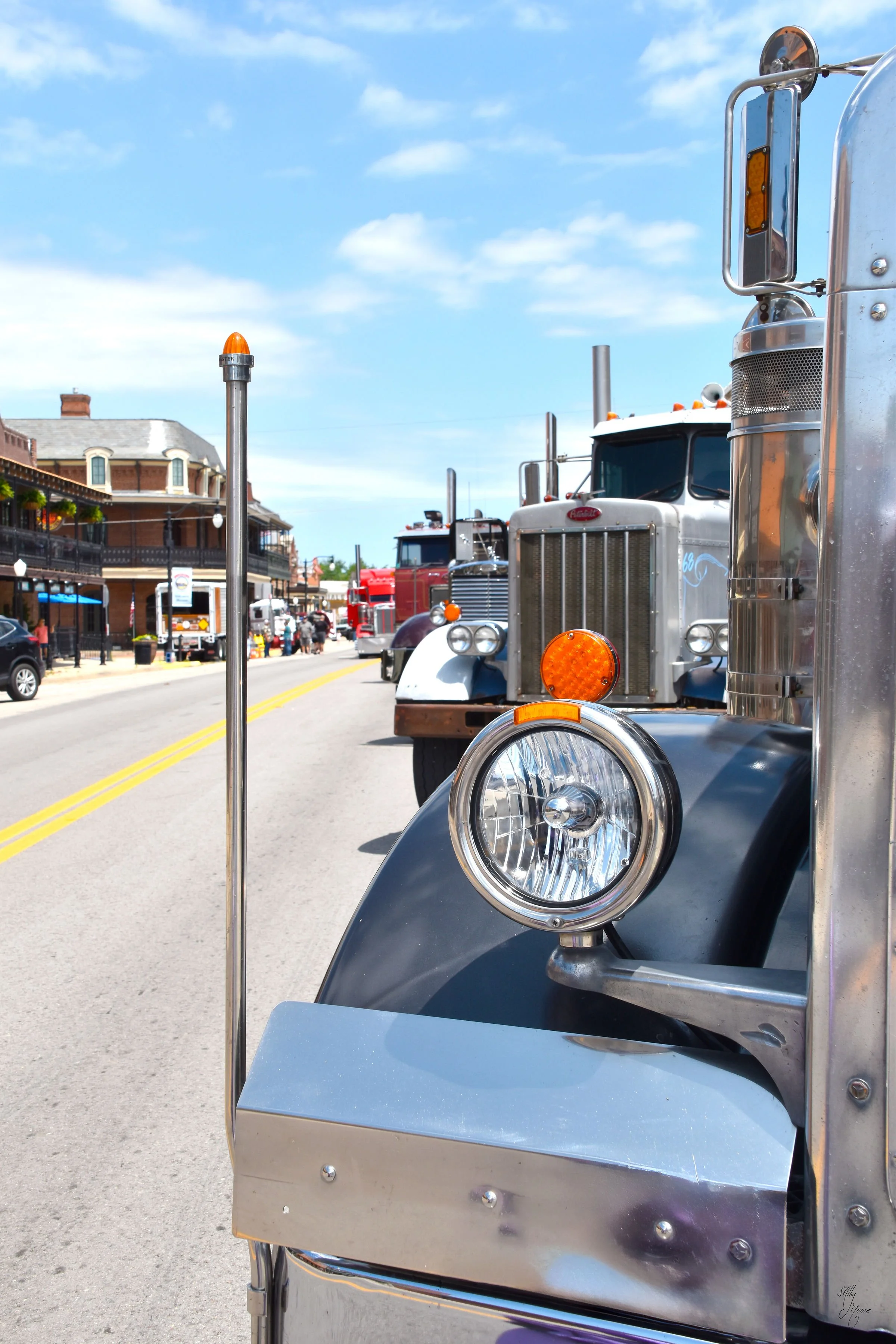 Close-up of the front of a vintage semi-truck with shiny metal details, parked on a street with other trucks and buildings in the background.