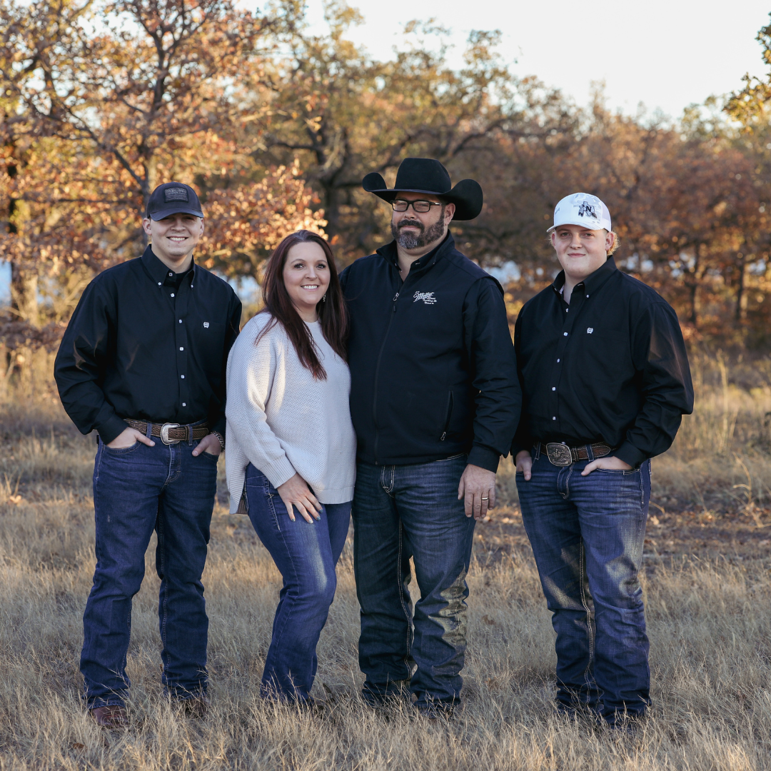 Four people standing outdoors in fall foliage, with two men wearing black shirts and one in a black cowboy hat, and a woman in a white sweater, smiling and posing for a family photo.