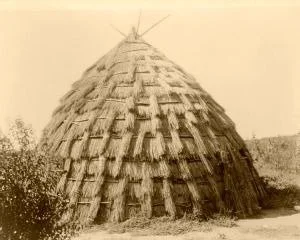 A large, dome-shaped structure made of woven or layered materials, resembling a wigwam or traditional hut, surrounded by trees or bushes.