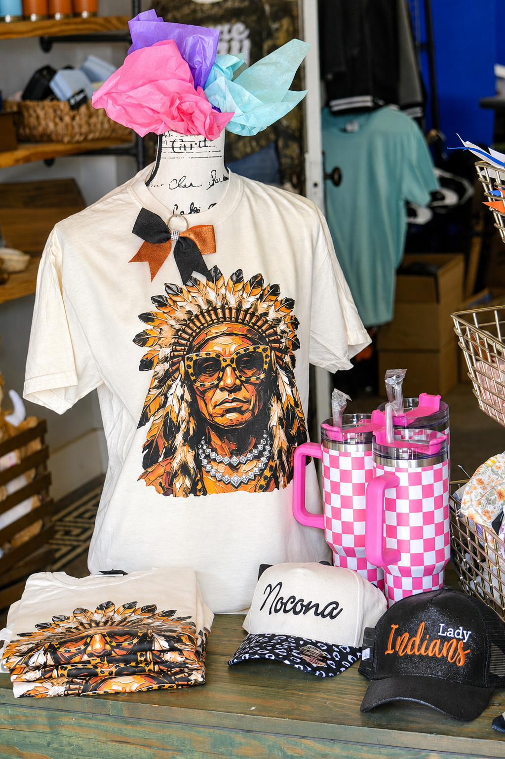 Display of T-shirts, hats, and drink tumblers at a market booth, featuring a Native American chief with sunglasses graphic, various caps, and pink and white checkered tumblers. Tissue paper decoration on a mannequin head. Nocona Texas