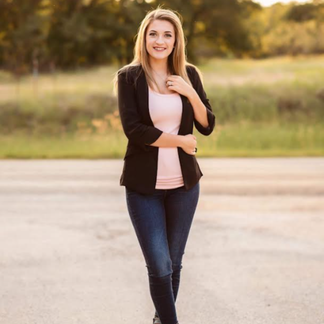 A young woman with blonde hair smiling, standing outdoors on a paved path with trees and greenery in the background, during golden hour.