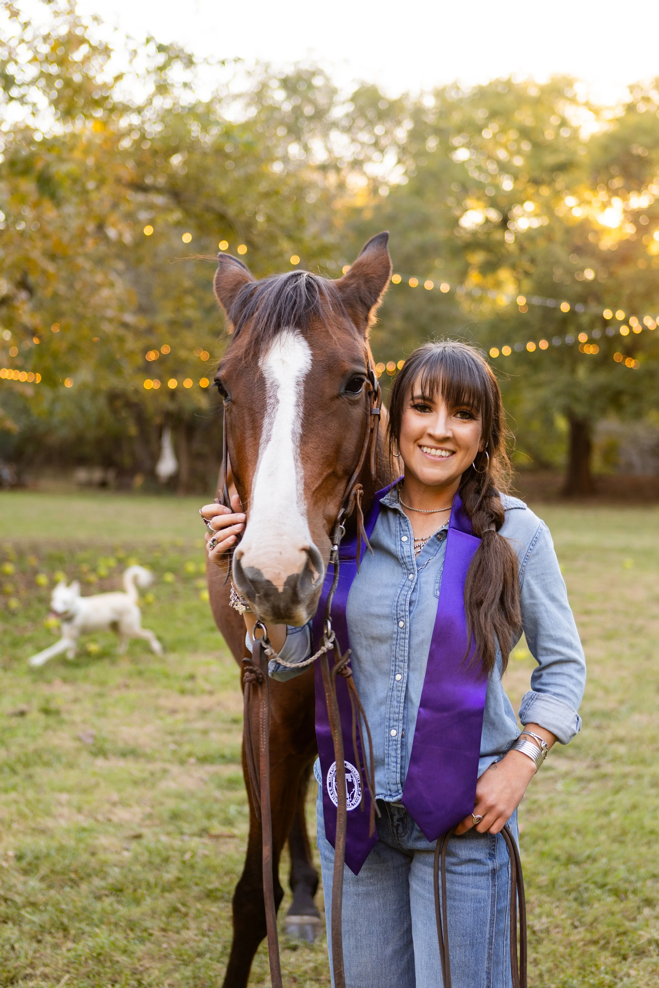 A young woman with long brown hair in a braid, wearing a denim shirt and purple stole, standing outside with a brown and white horse during sunset, smiling. A small white dog is running in the background.