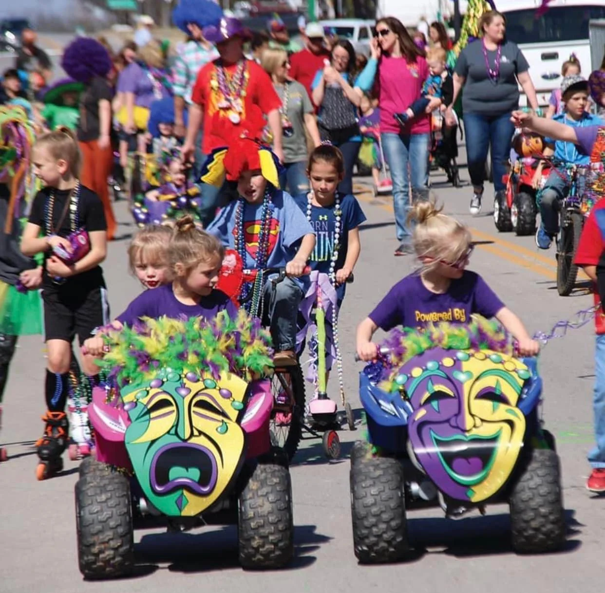Children and adults participating in a colorful parade on a street, with kids riding decorated toy vehicles featuring theatrical masks, some wearing costumes and beads, and a crowd of spectators and other participants in the background.