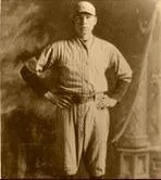 A man standing indoors near a column, wearing a flat cap, plaid shirt, and suspenders, with his hands on his hips.