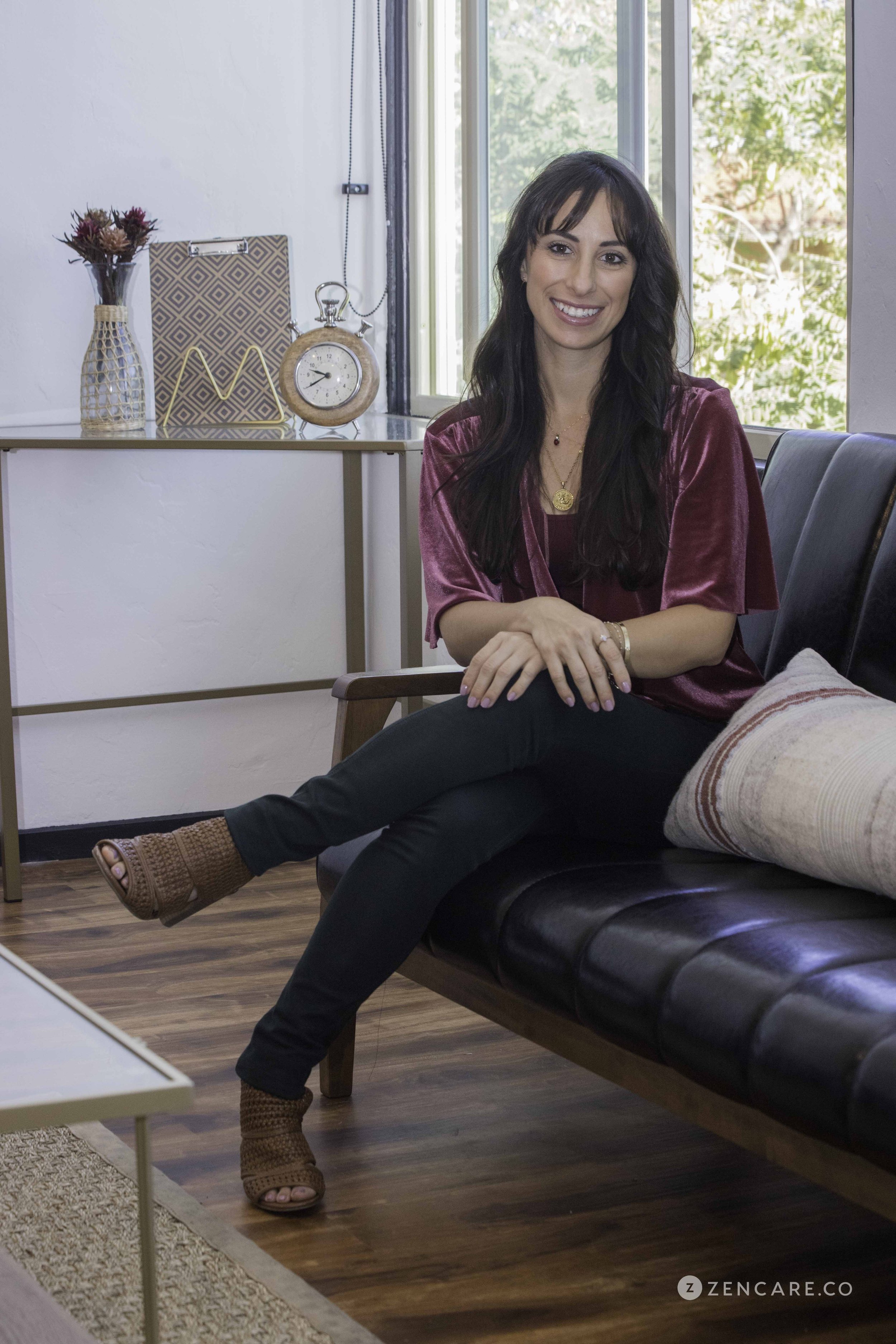 A portrait of LMFT Diandra Medina who is a holistic therapist in San Diego with long brown hair who is wearing dark green pants and an auburn shirt and sitting in an office on a black couch with a clock, flowers, and notebook in the background.