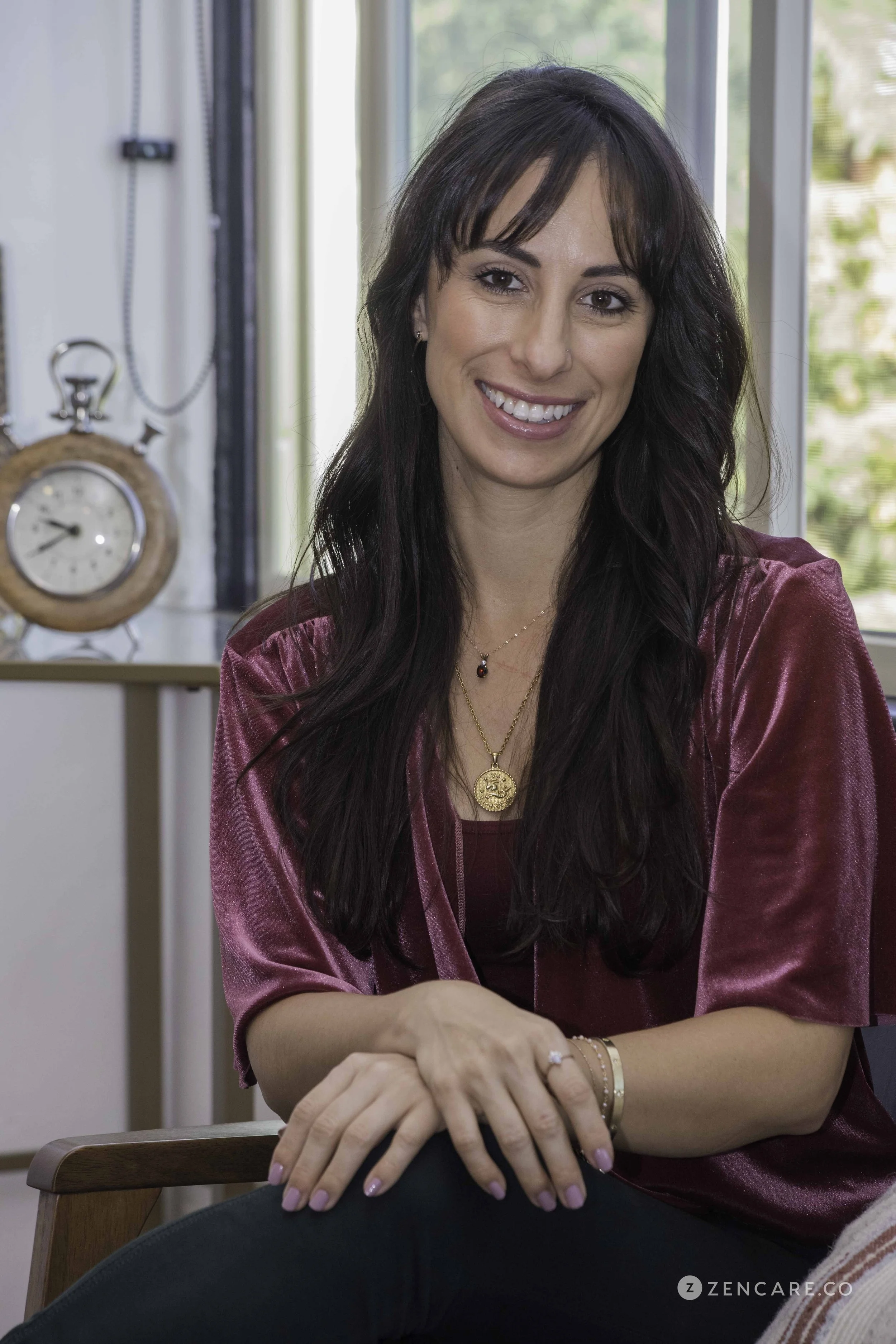 LMFT Diandra Medina who does trauma therapy CA and has long brown hair and is wearing gold jewelry wearing and a maroon top is sitting in front of a clock and smiling toward the camera.