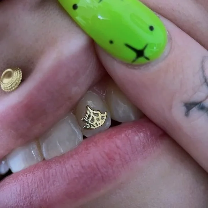Close-up of a person's slightly open mouth showing a metallic tooth jewel with a spiderweb design, a gold nose ring, and a brightly painted green fingernail with a bear face design.