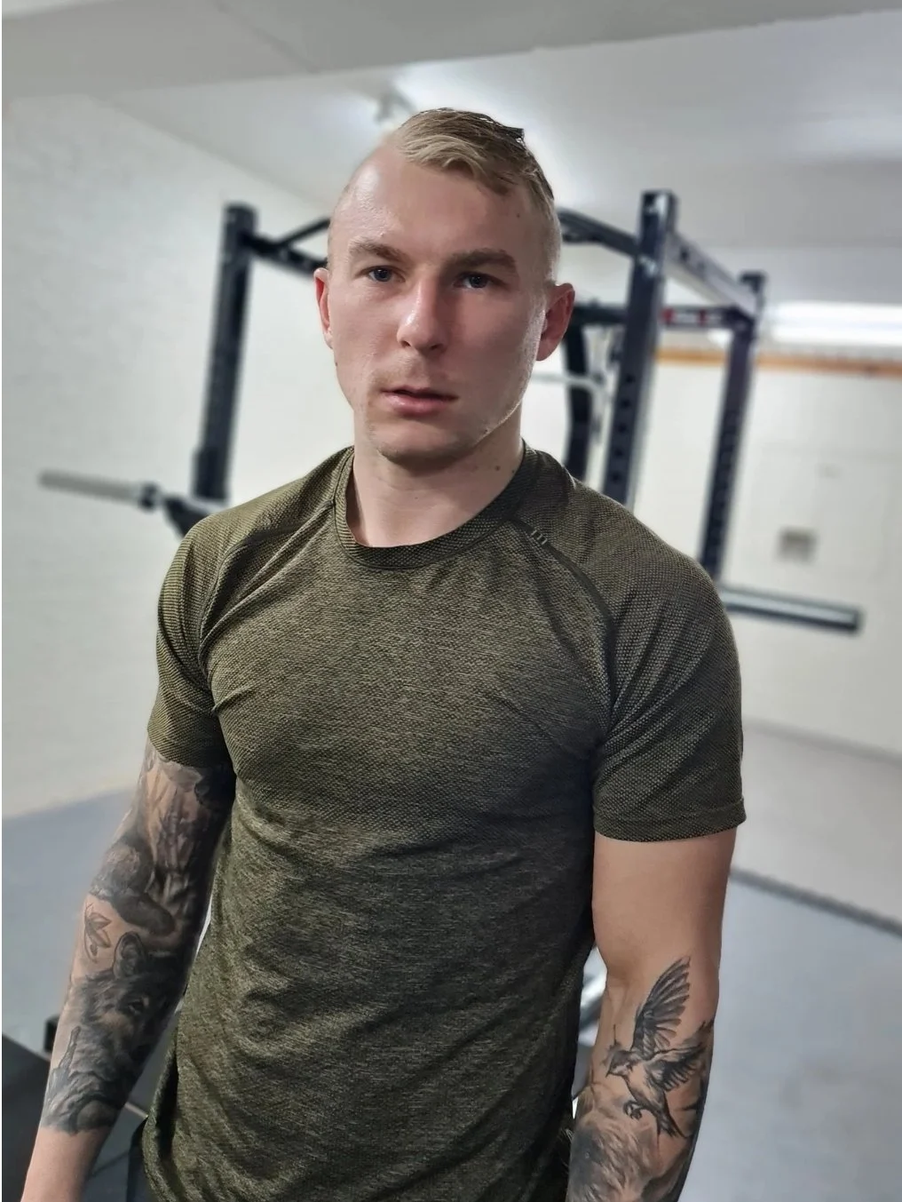 A young man with tattoos on his arms wearing an olive-green athletic shirt, standing in a gym with equipment in the background.