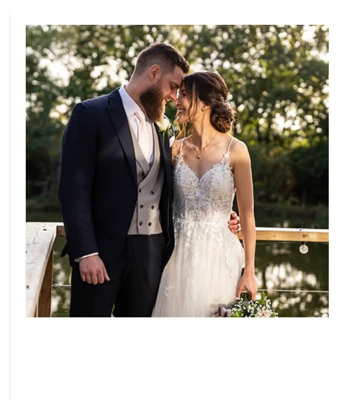 A couple stand facing each other, smiling, with their foreheads touching. The bride is wearing a white tulle dress with spaghetti straps and is holding a bouquet by her side. The groom has his arm around her and the sun beams down on them