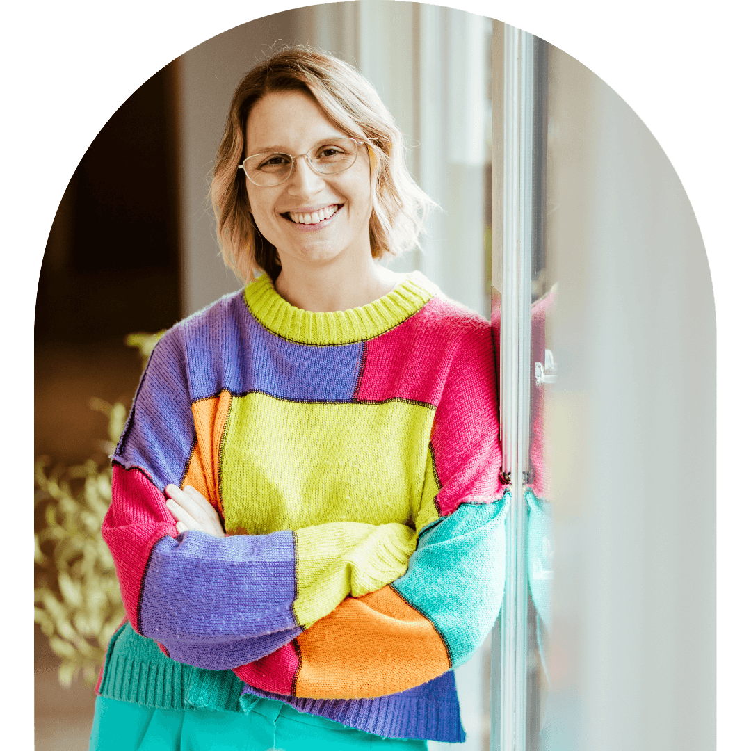Image of Hollie leaning on a window with her arms crossed, smiling at the camera. She has blonde hair, in a wavy style, rounded rectangular silver glasses and is wearing a patchwork pink, purple, orange, green and teal jumper with teal trousers