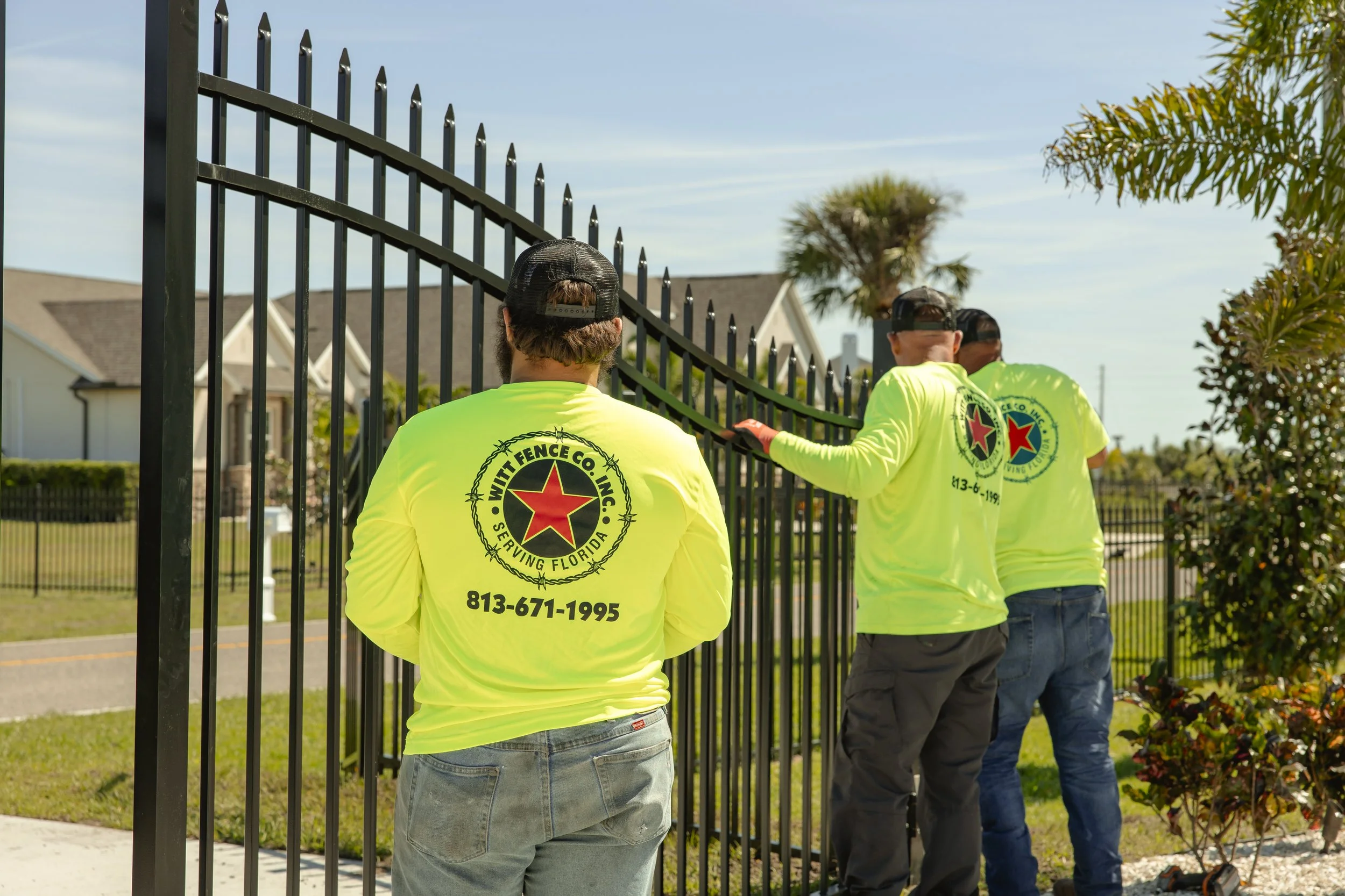 Four workers installing or repairing a black metal fence in a residential neighborhood. They are wearing bright yellow shirts with a red star logo and contact information on the back. One worker is facing away, while the other three are working on or near the fence.