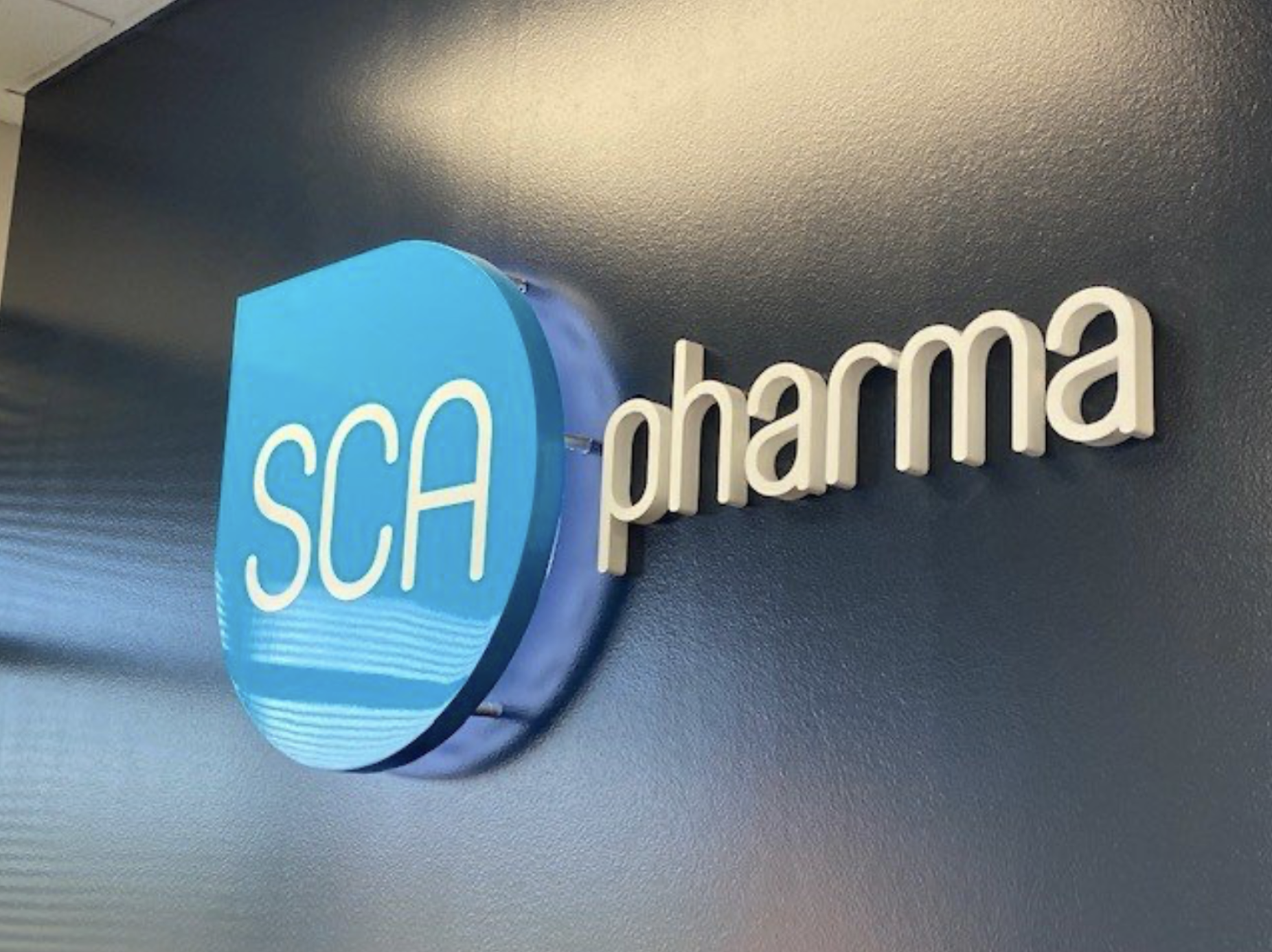 A wall-mounted sign with a blue circular logo containing the words "SCA" and the word "pharma" next to it, on a dark blue wall.