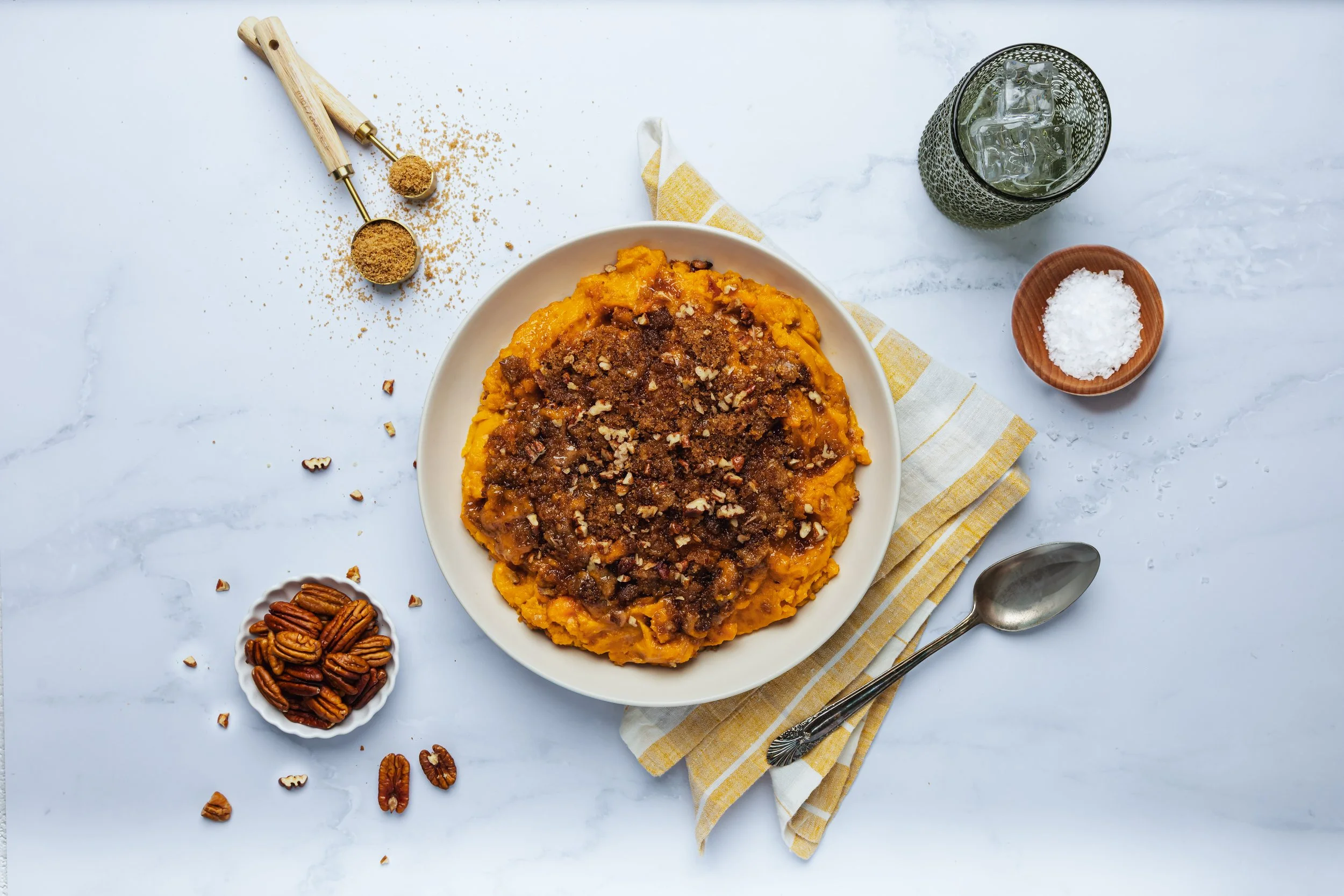 A plate of sweet potato casserole topped with brown sugar and chopped pecans, with a bowl of pecans, coarse salt, and a glass of ice water around it.