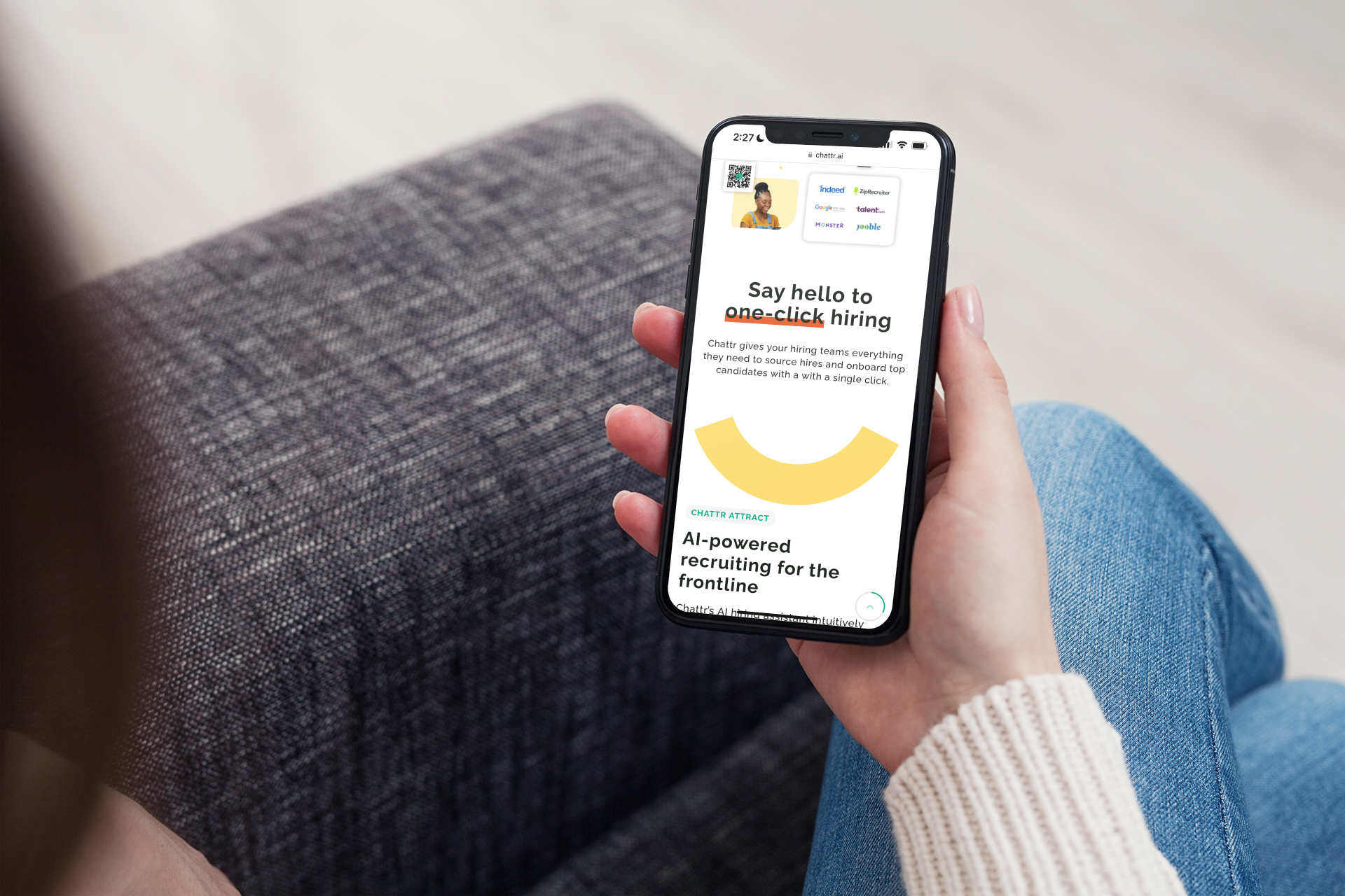 Person holding a smartphone with a website about AI-powered recruiting on the screen, sitting on a gray textured couch, wearing blue jeans and a light-colored sweater.