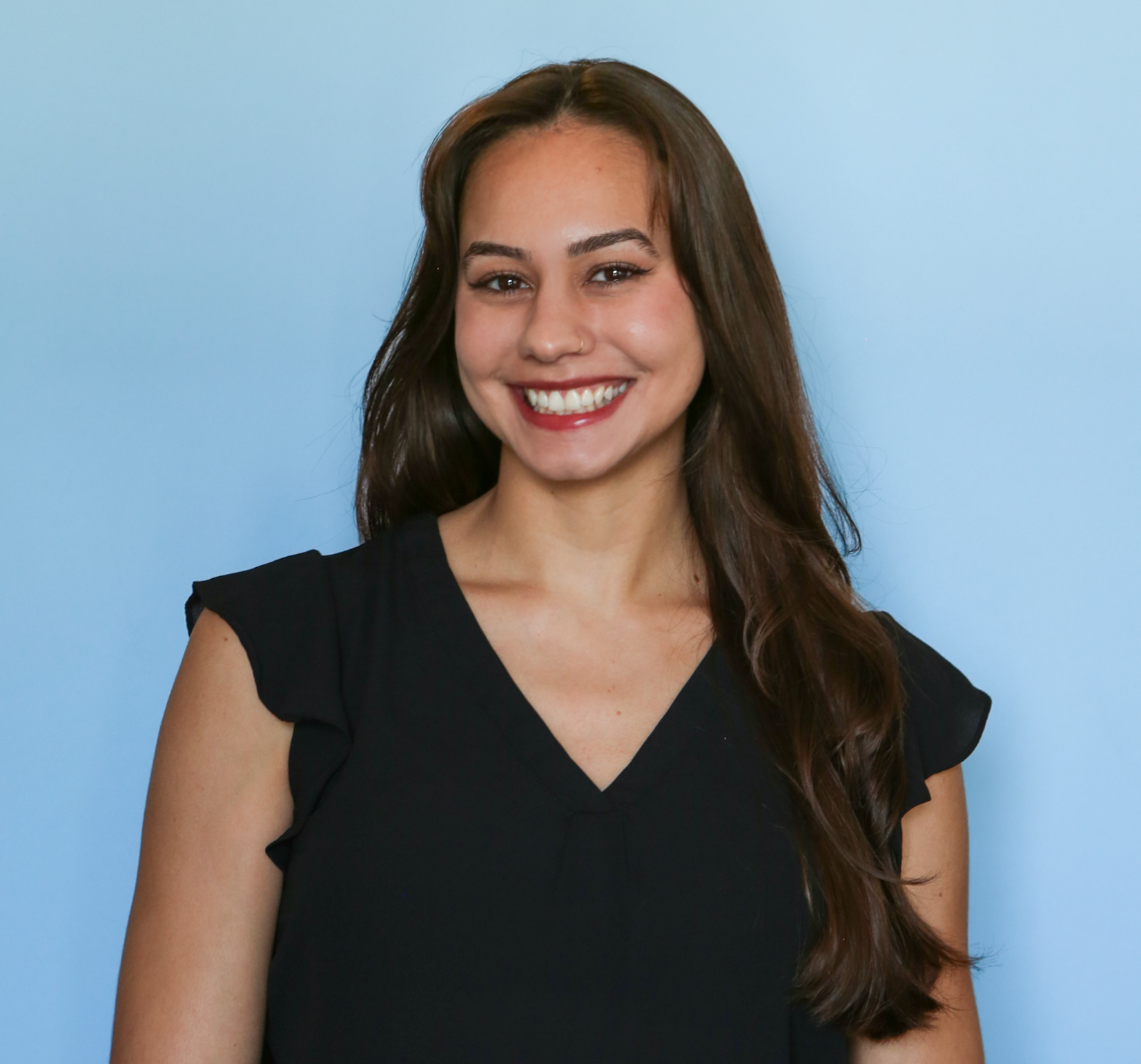 A young woman with long brown hair smiling at the camera, wearing a black sleeveless top, against a light blue background.