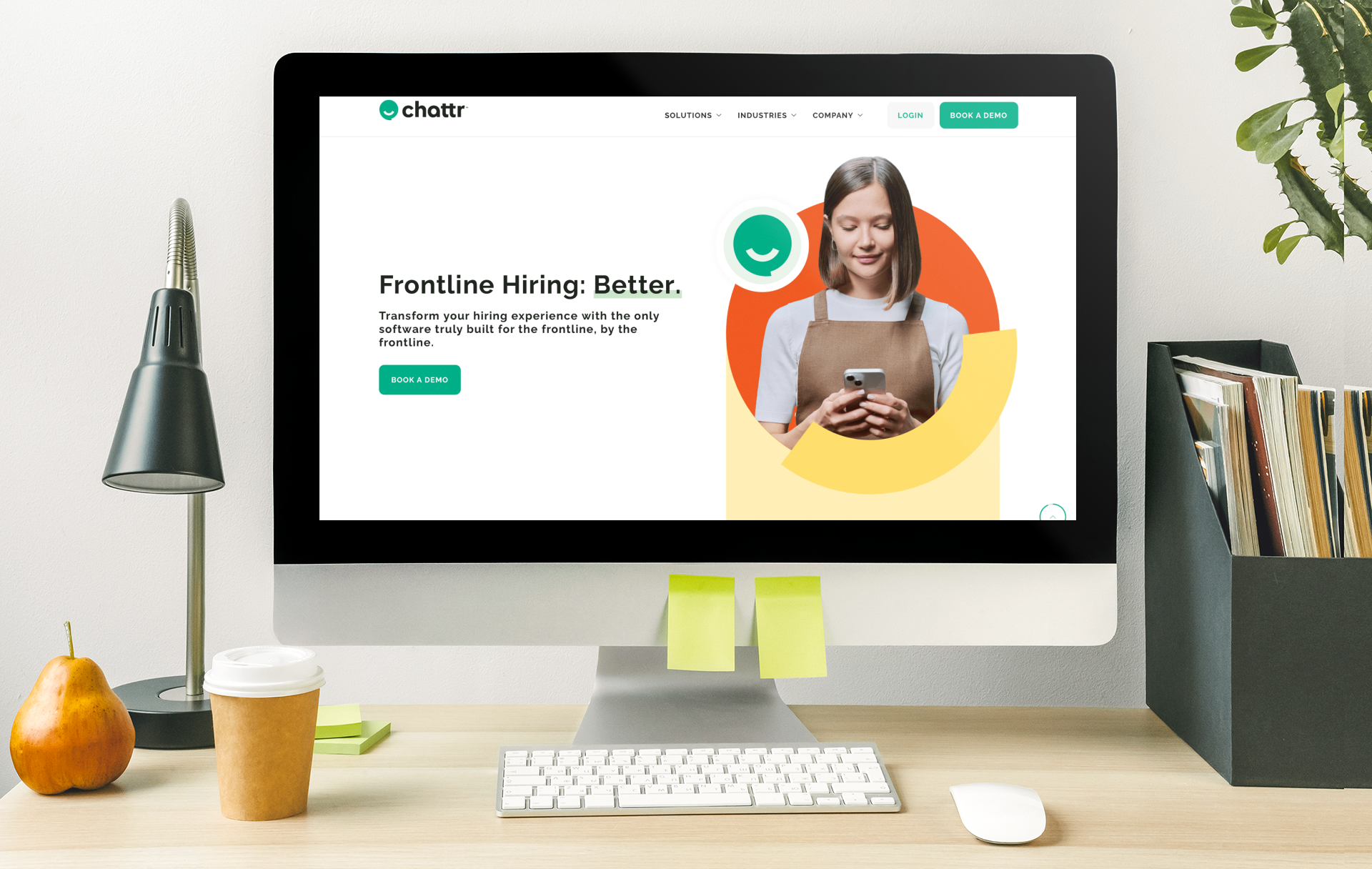 Computer screen displaying a website for Chattr with a woman holding a phone, the website has the headline "Frontline Hiring: Better" with a green "Book a Demo" button. The desk has a pear, a coffee cup, a keyboard, a mouse, and a black file organizer with books. A desk lamp and a green plant are also visible.