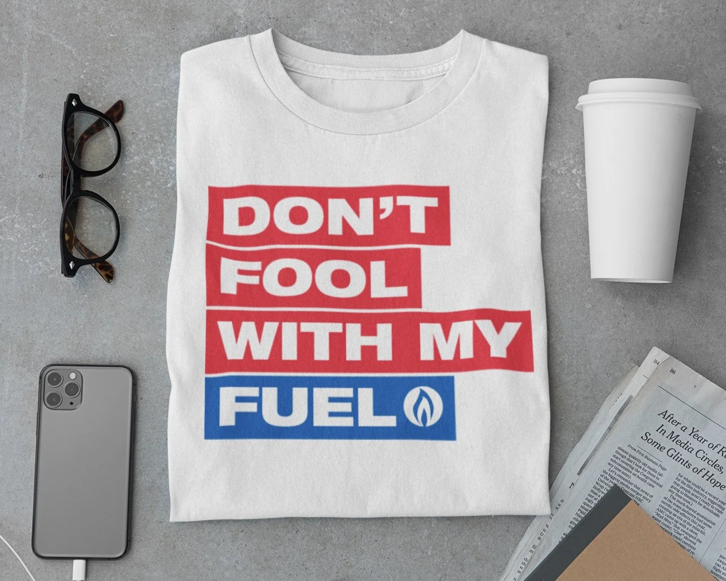 White T-shirt with the text 'DON'T FOOL WITH MY FUEL' in red and blue letters, with a fuel flame icon, lying on a grey surface surrounded by glasses, a smartphone, a paper coffee cup, and newspapers.