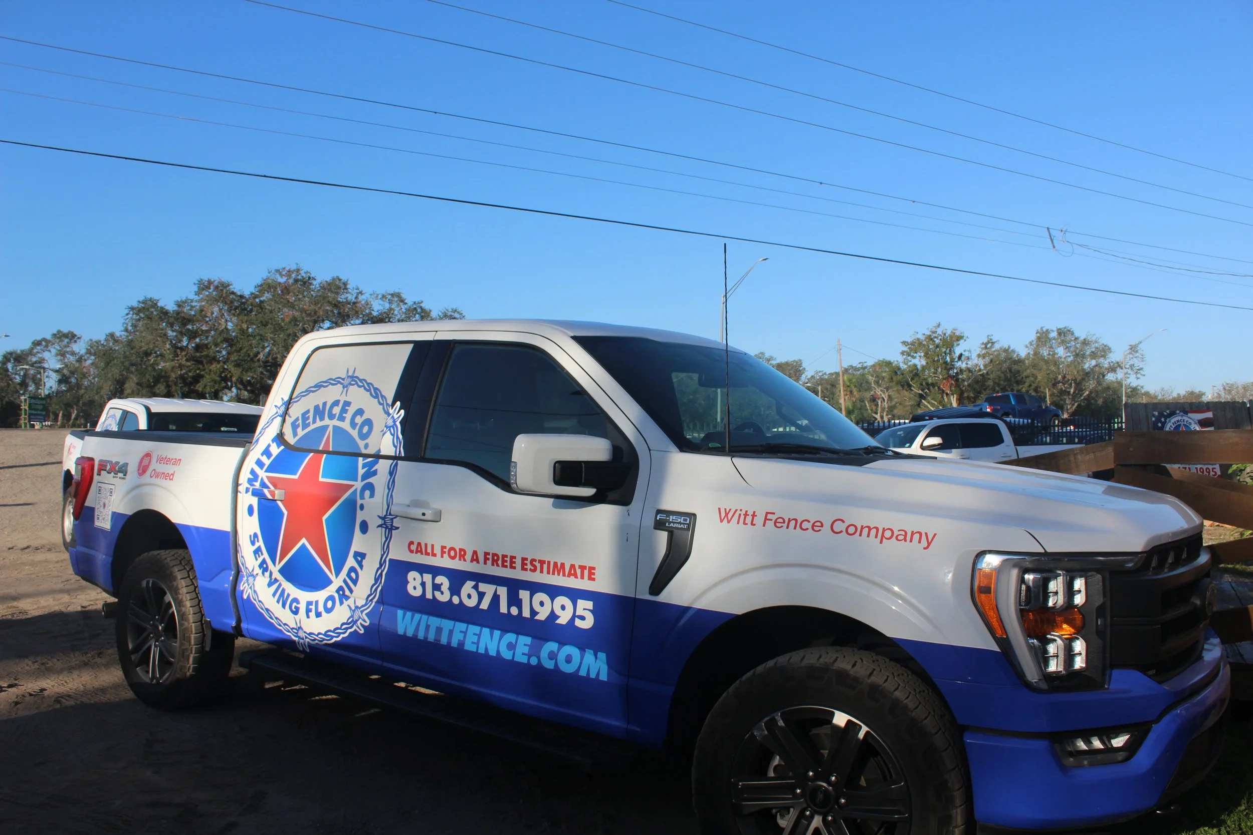 A white and blue pickup truck with the logo of Witt Fence Company. The truck displays contact information in red and blue text and is parked outdoors near a wooden fence with trees and blue sky in the background.