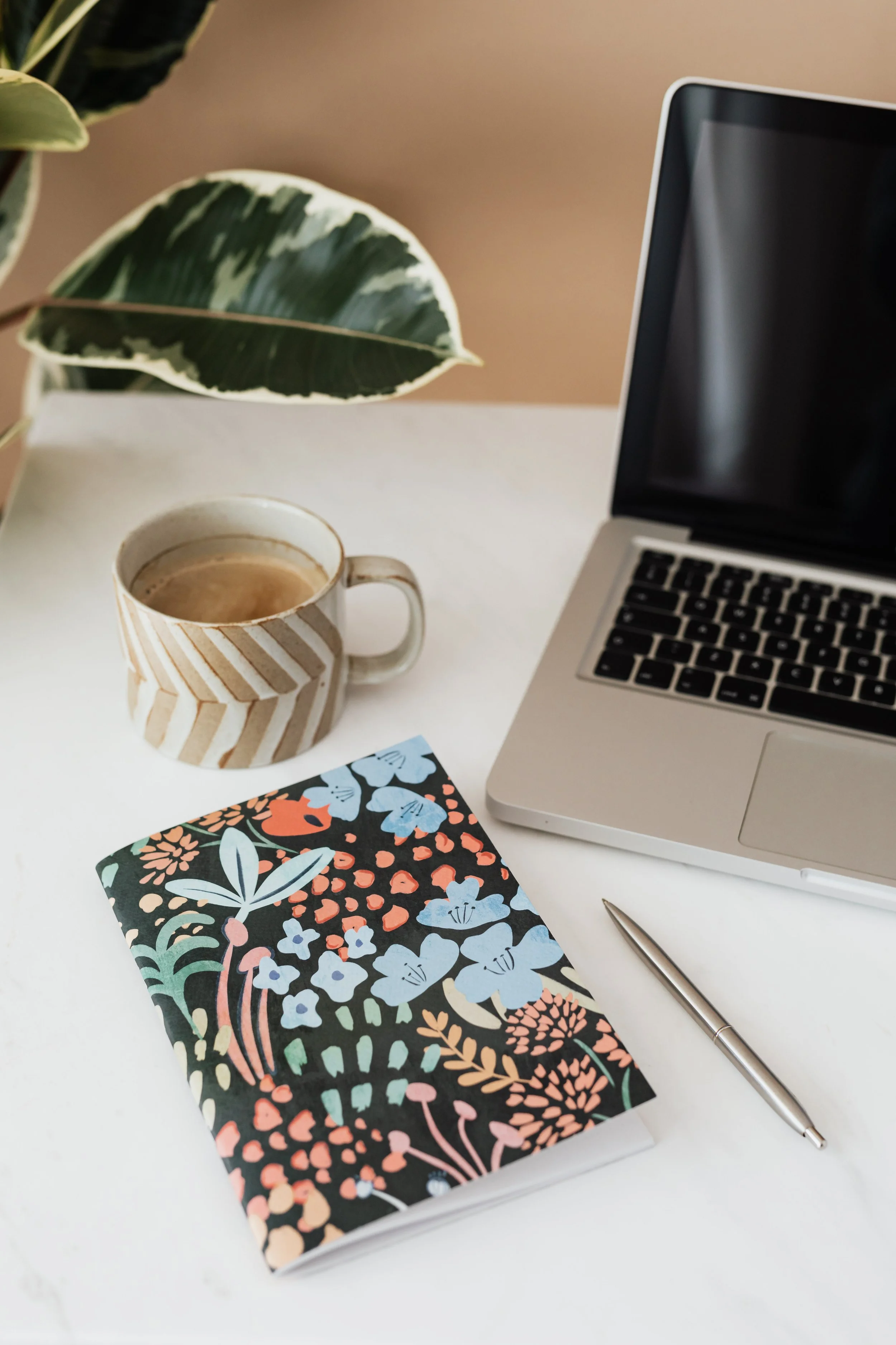 Uma mesa branca com um caderno com estampa floral, uma caneta prateada, uma caneca de café com listras bege e brancas, um laptop parcialmente aberto, uma planta com folhas variegadas de verde e branco e um cartão ou folha de papel azul.
