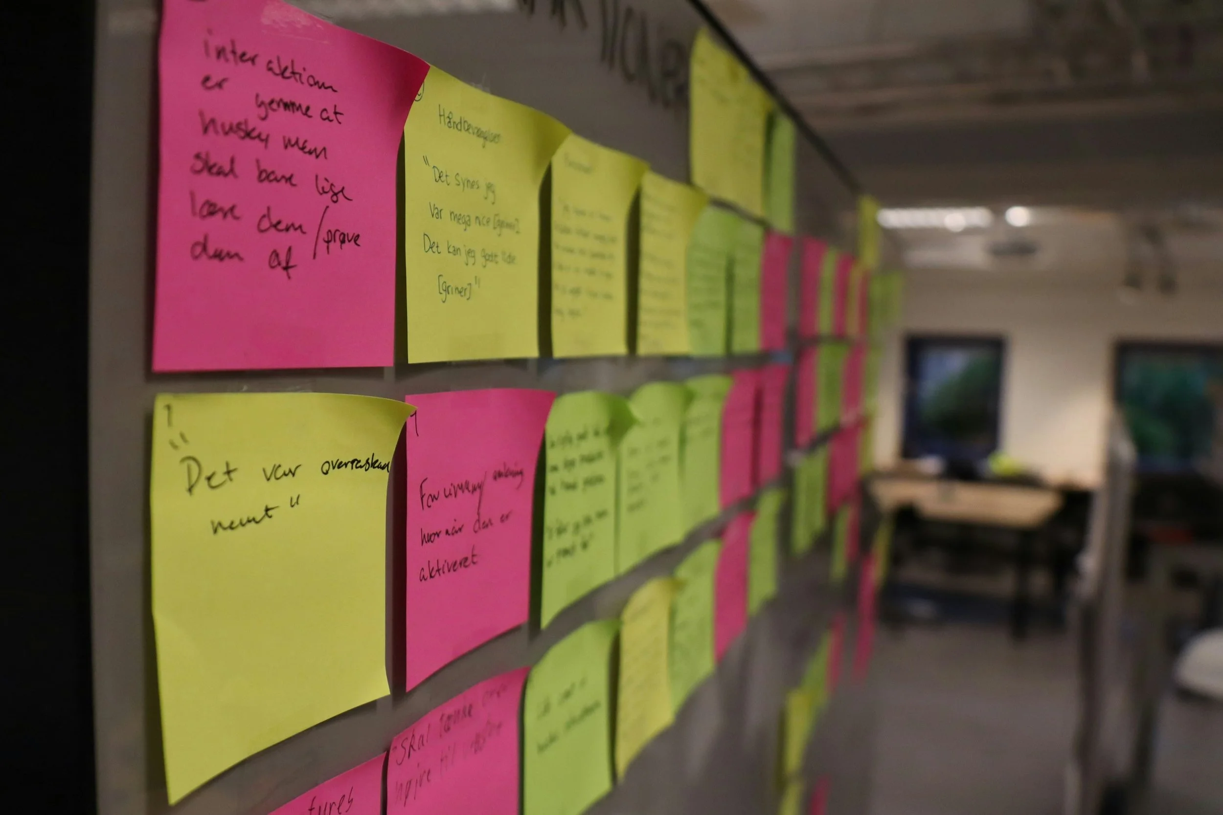 Close-up de uma parede com post-its rosa e amarelos dispostos em fileiras, com anotações escritas à mão em cada um deles.