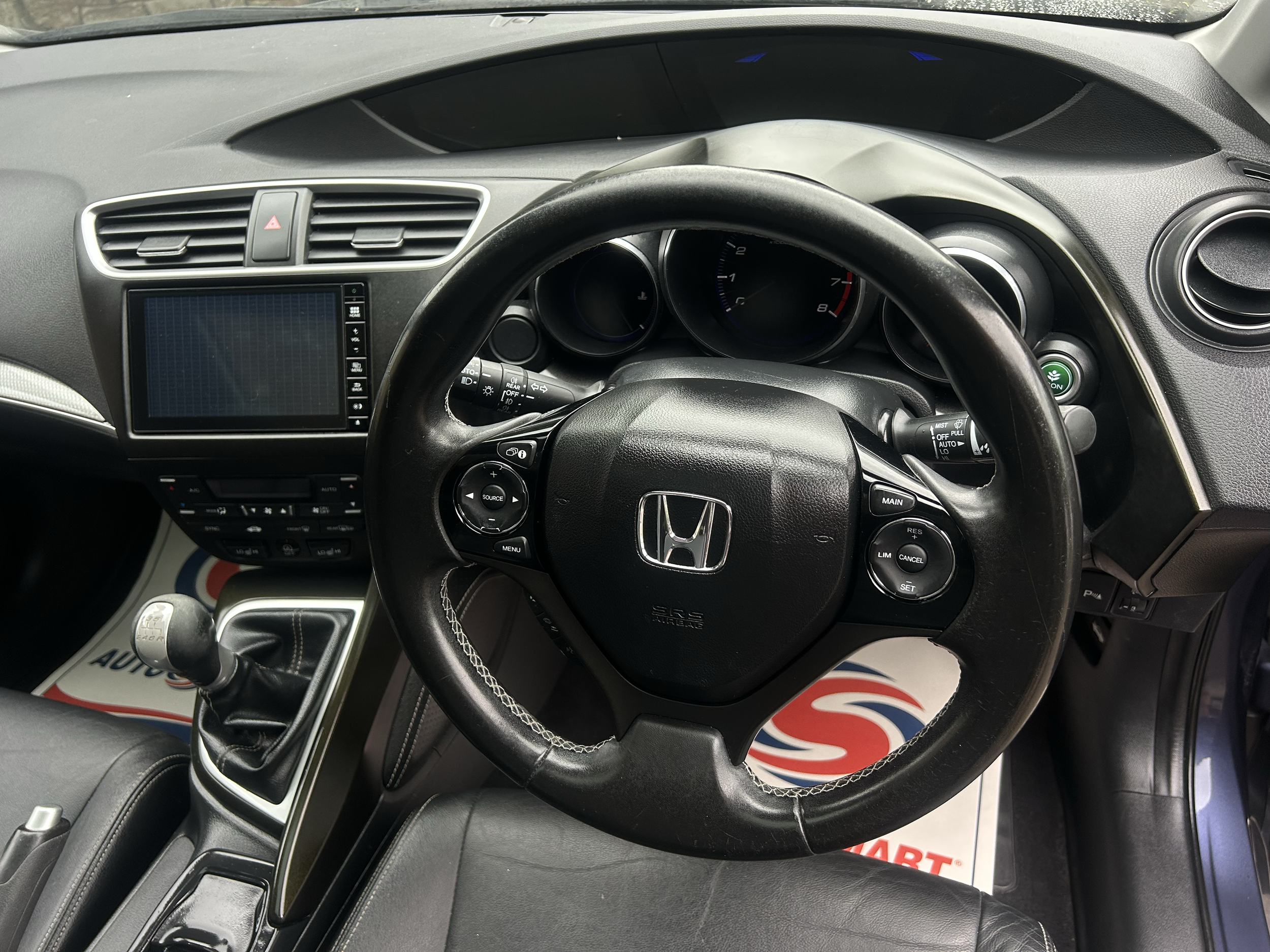 The interior of a Honda car, focusing on the steering wheel, dashboard, and center console. The steering wheel has various control buttons, and the dashboard includes an analog speedometer and tachometer. The center console features an infotainment s