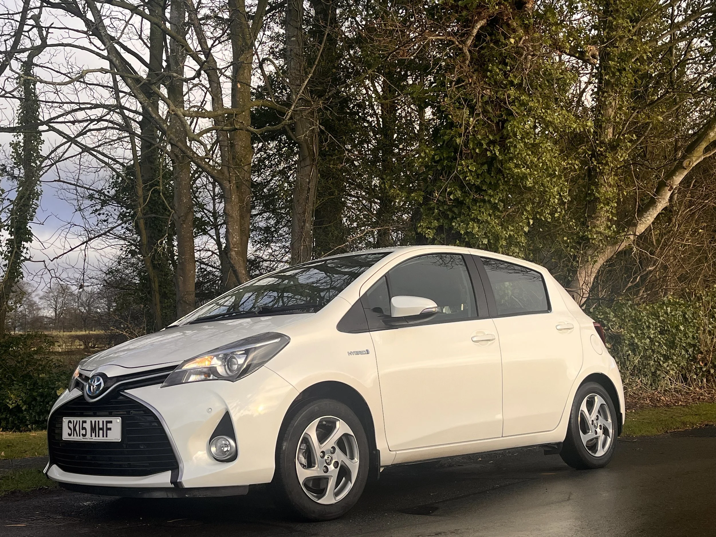 A white hatchback Toyota car parked on the side of the road, with trees and bushes in the background during daytime.