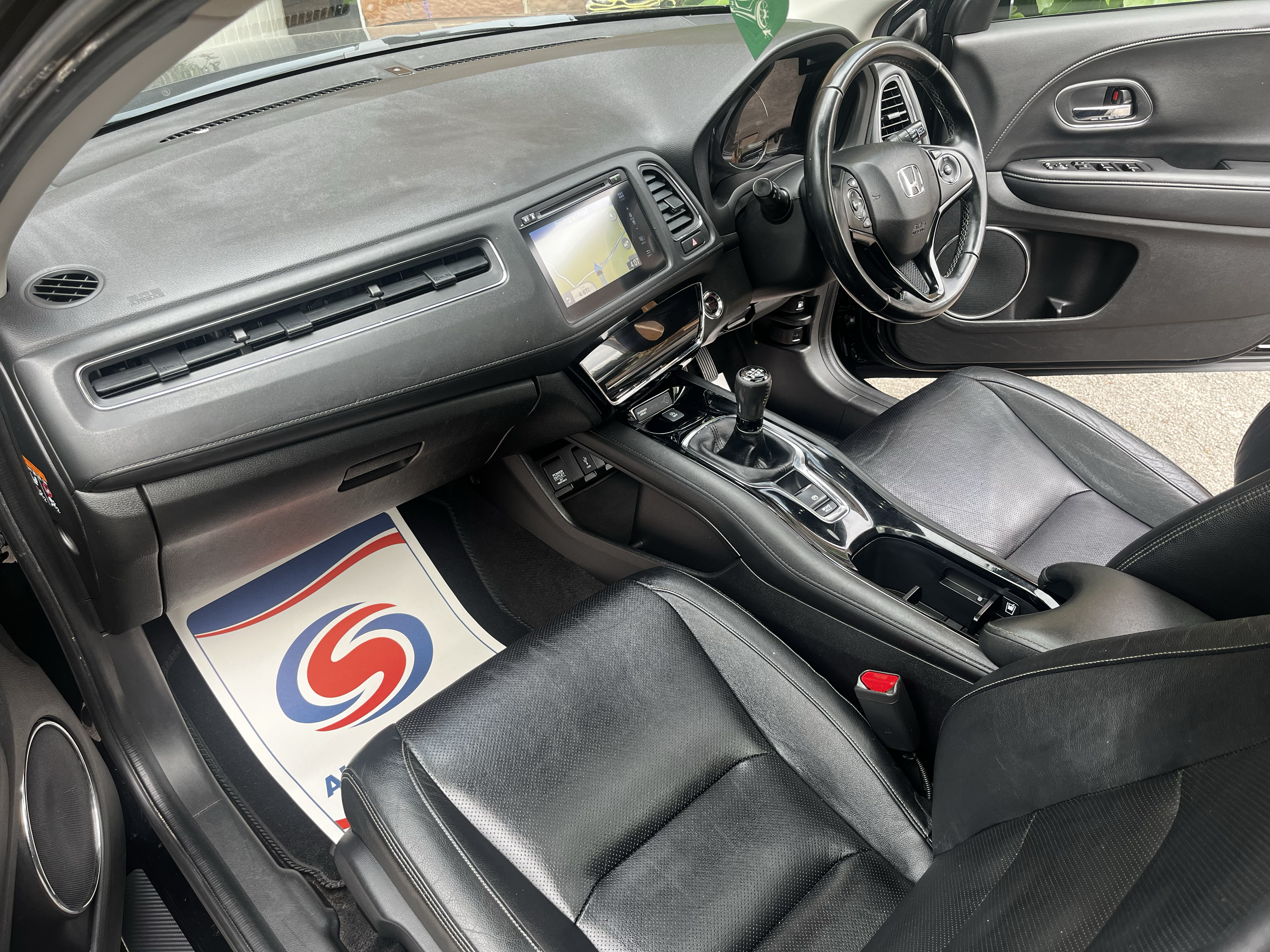 Interior view of a car dashboard showing a GPS navigation screen, steering wheel with Honda logo, gear shift, and black leather seats.