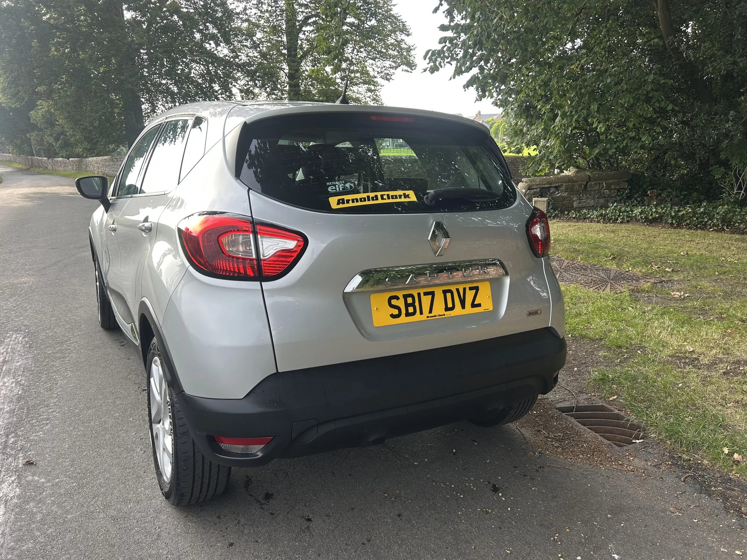 Rear view of a silver Renault Captur parked on a street with a yellow UK license plate reading 'SB17 DVZ' and decorative decal stickers on the rear window.