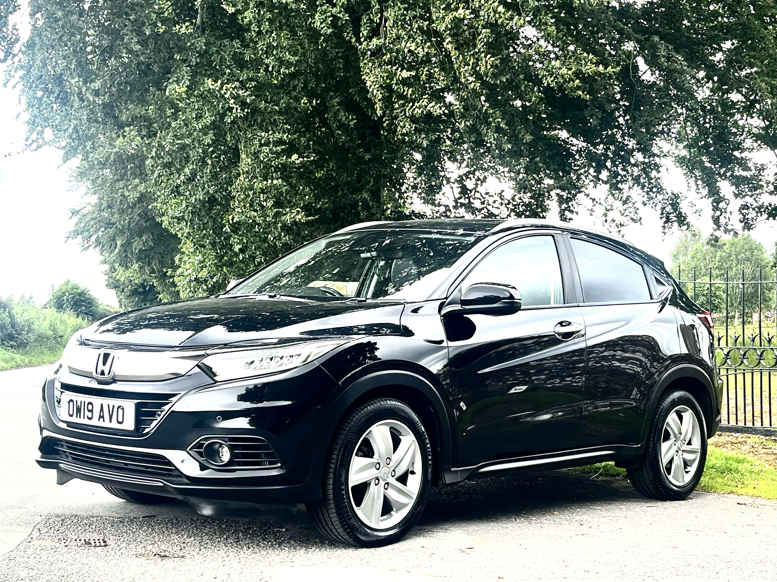 A black Honda Civic parked outdoors on a paved road, with large green trees and an iron fence in the background, under a bright, partly cloudy sky.