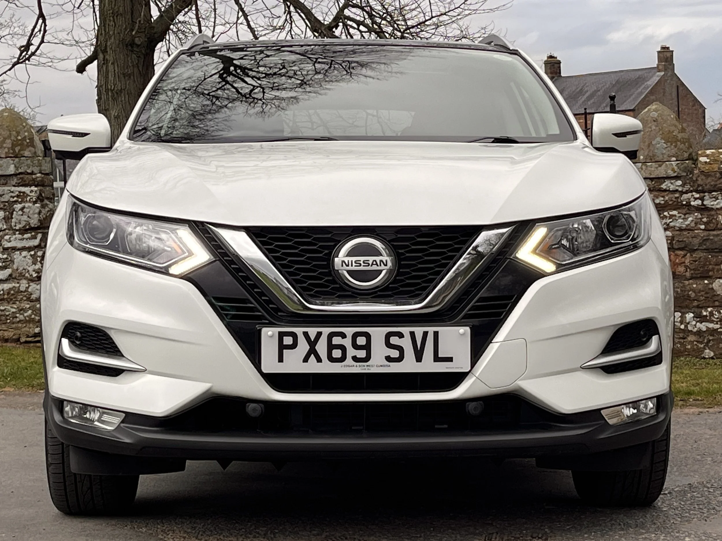 Front view of a white Nissan SUV parked outdoors, with stone wall and trees in the background.