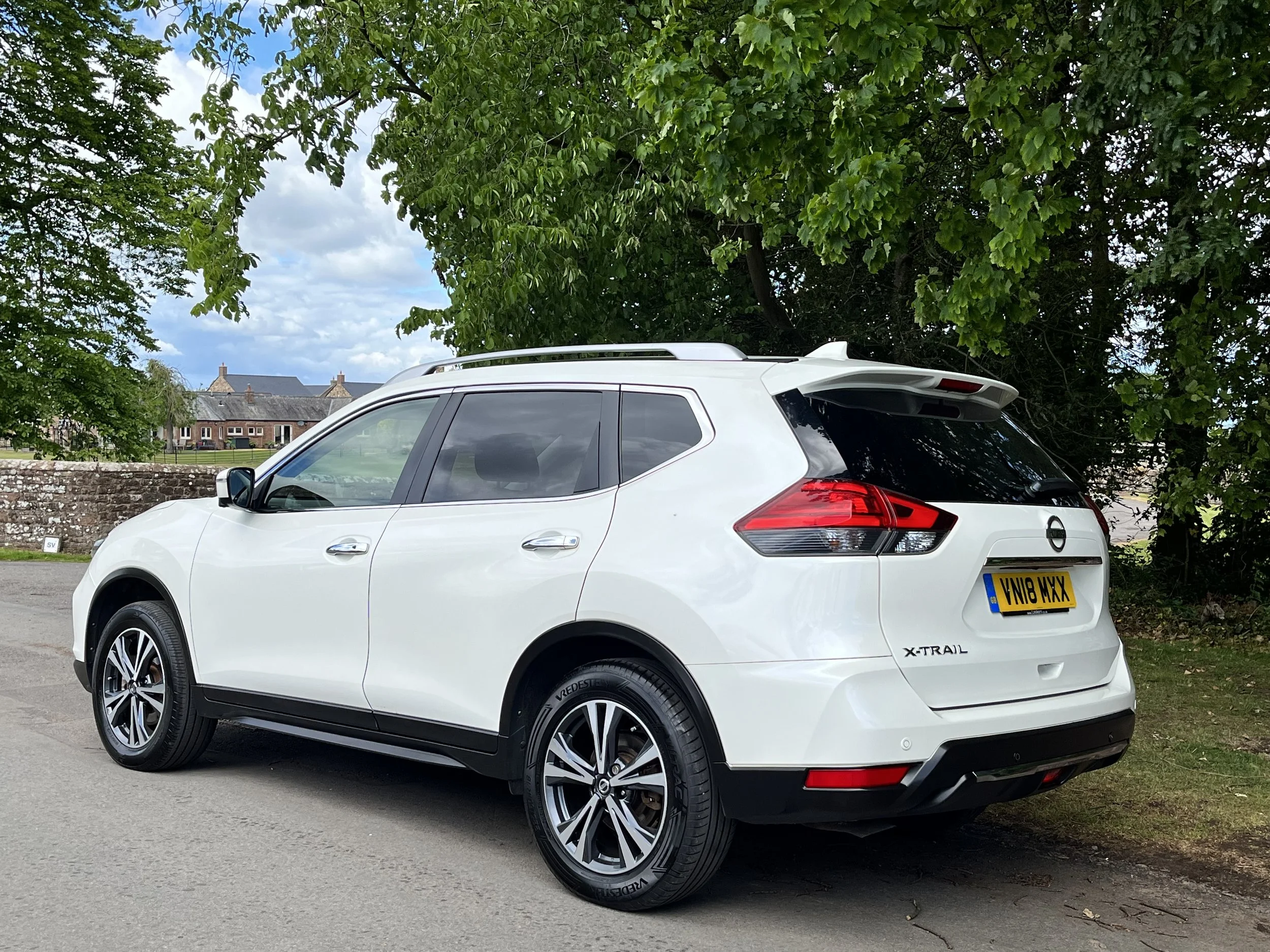 White Nissan X-Trail SUV parked on the side of a road, with green trees overhead and a brick building in the background.