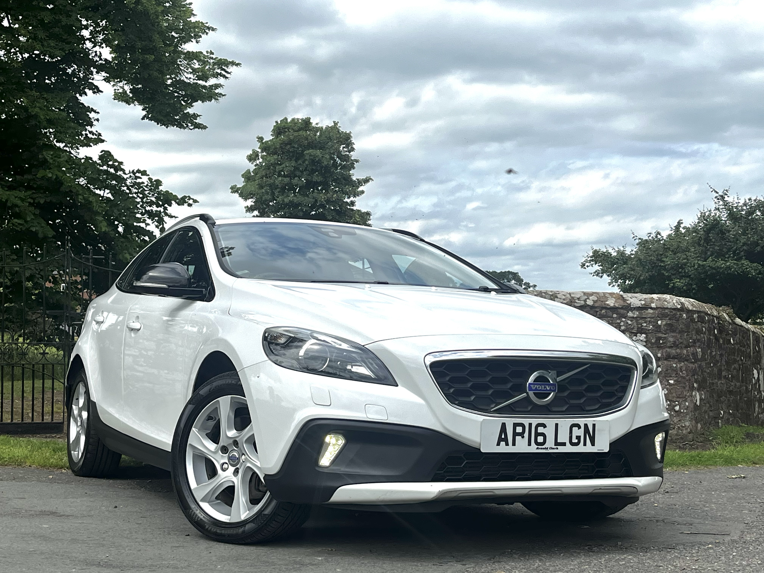 A white Volvo car parked outdoors on a cloudy day. The car has a black grille, is facing forward, and displays a UK license plate reading 'AR16 LGN.' There are green trees and a stone wall in the background.