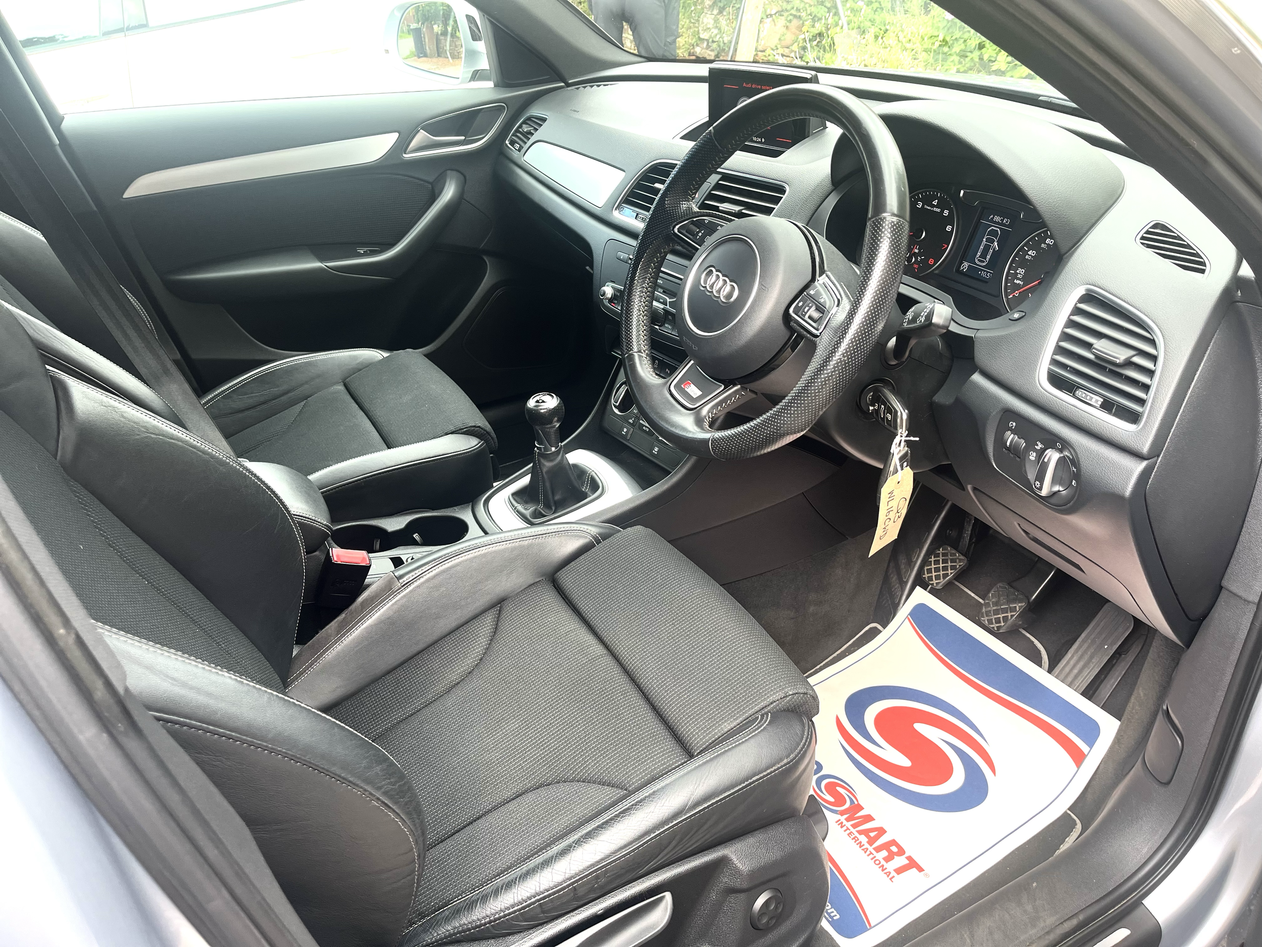 The interior of a car shows a black leather seat, a steering wheel with the Audi logo, and dashboard instruments, with a gear shift lever in the center console.