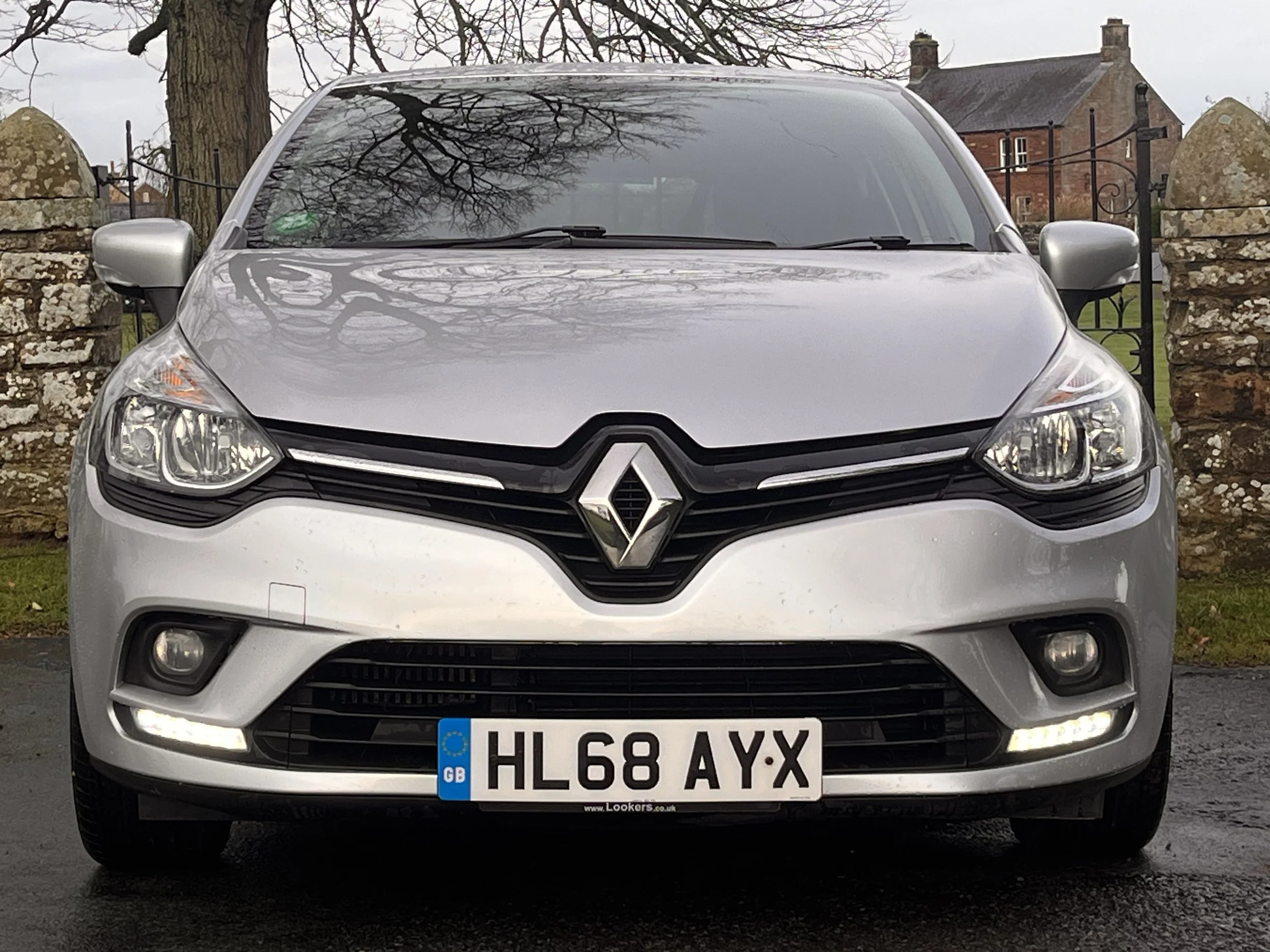 Front view of a silver Renault car parked outdoors with trees and an old house in the background.